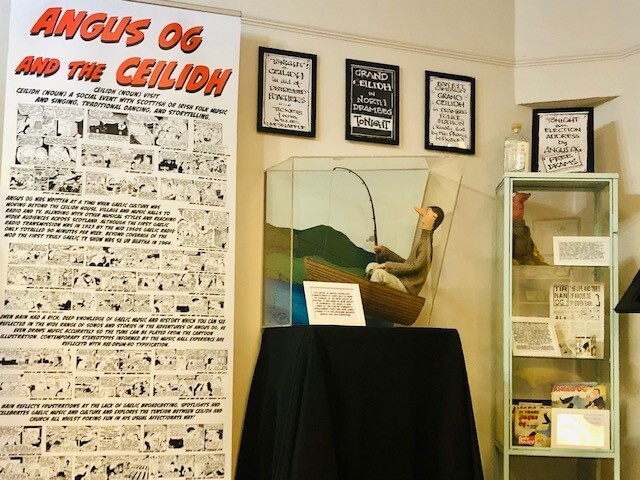 An exhibition of Angus Og items including a full pop up banner of comic strips, a small Angus Og figurine in a boat with a fishing road and various other framed items on the walls behind.