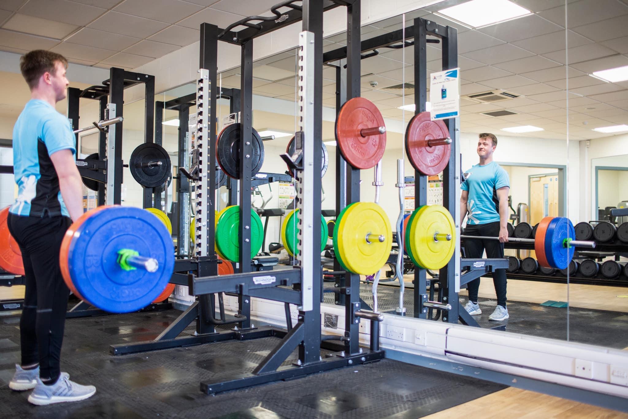 A person lifts a barbell with colourful weight plates in a well‑equipped gym, standing in front of a squat rack and a mirrored wall.
