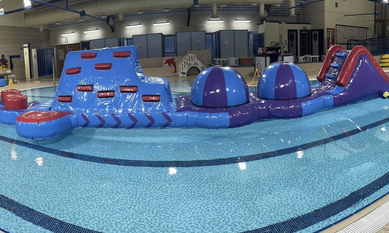A large inflatable obstacle course floating in a swimming pool inside an indoor leisure centre.