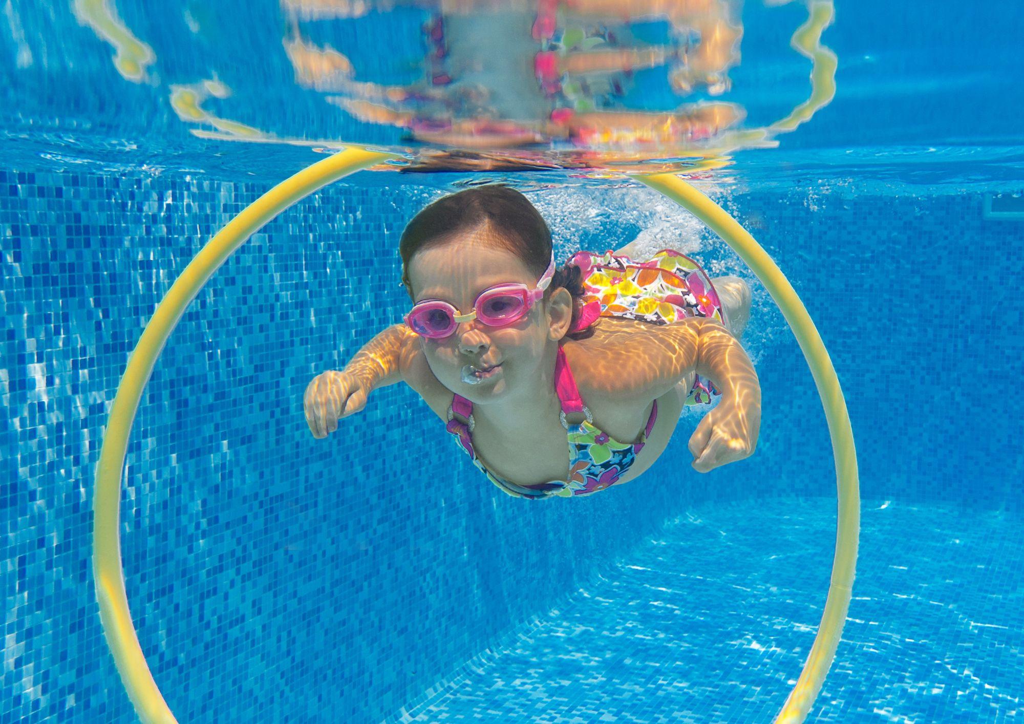 A child in a colorful swimsuit swims underwater through a circular hoop in a pool with blue tiled walls.