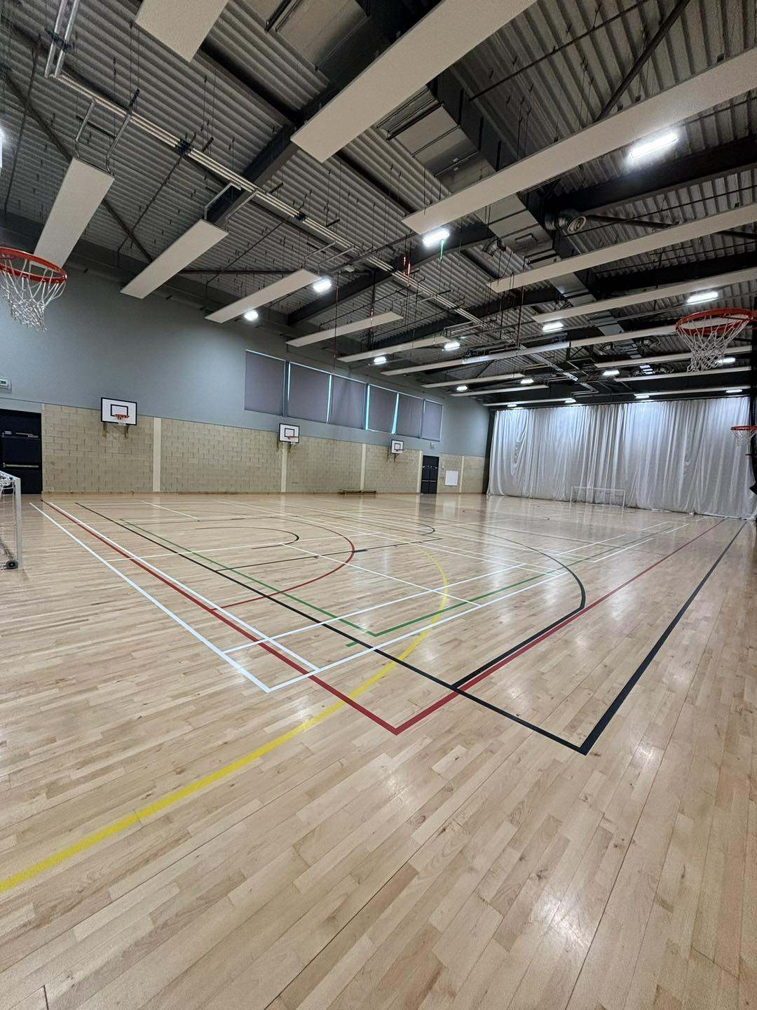 Large indoor sports hall with a wooden floor, multiple court markings for different sports, and basketball hoops along the walls.