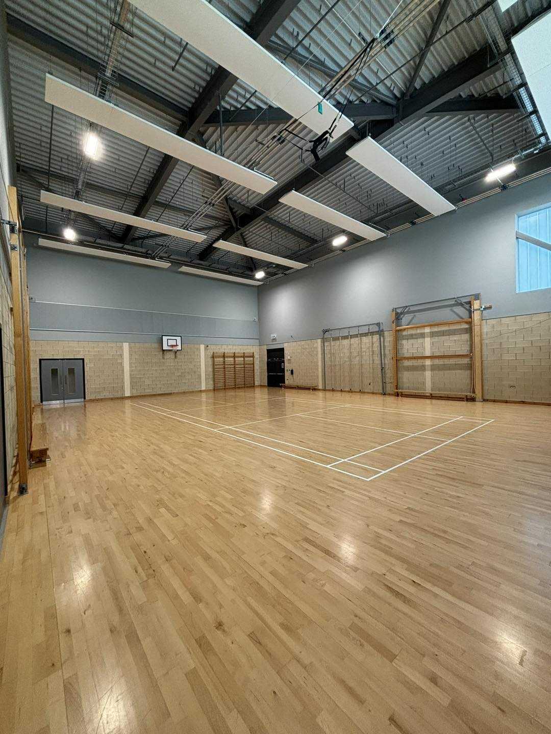 Empty indoor sports hall with a polished wooden floor, marked for badminton, and a basketball hoop on the far wall.