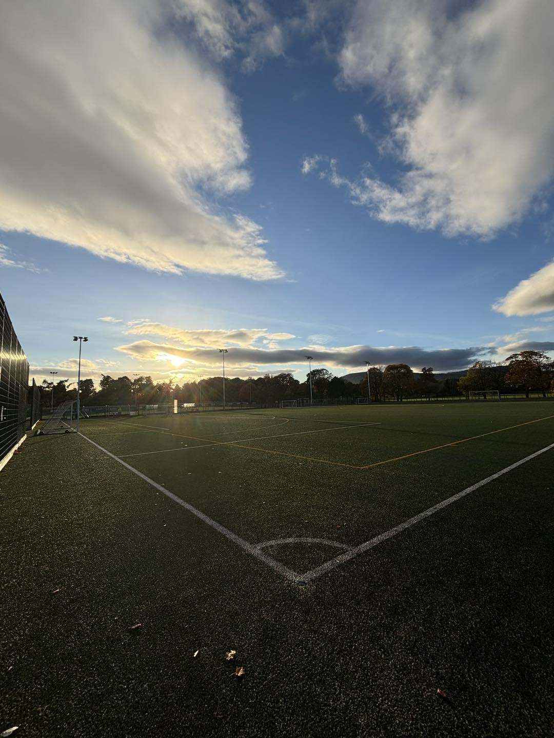 Outdoor sports pitch with artificial turf under a bright blue sky and scattered clouds, with the sun setting in the background.