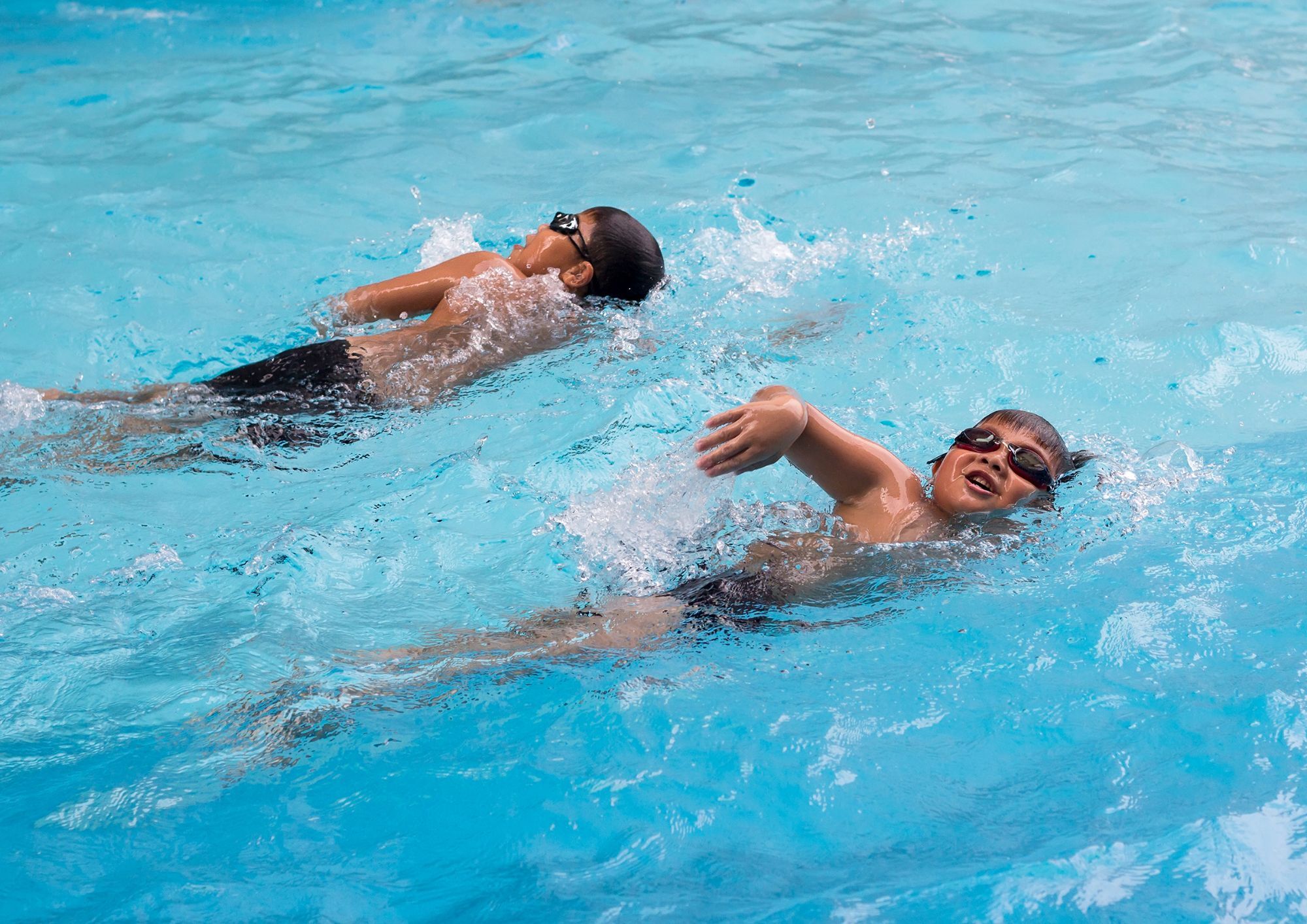 Two children swim front crawl in a bright blue pool, creating splashes as they move forward side by side.