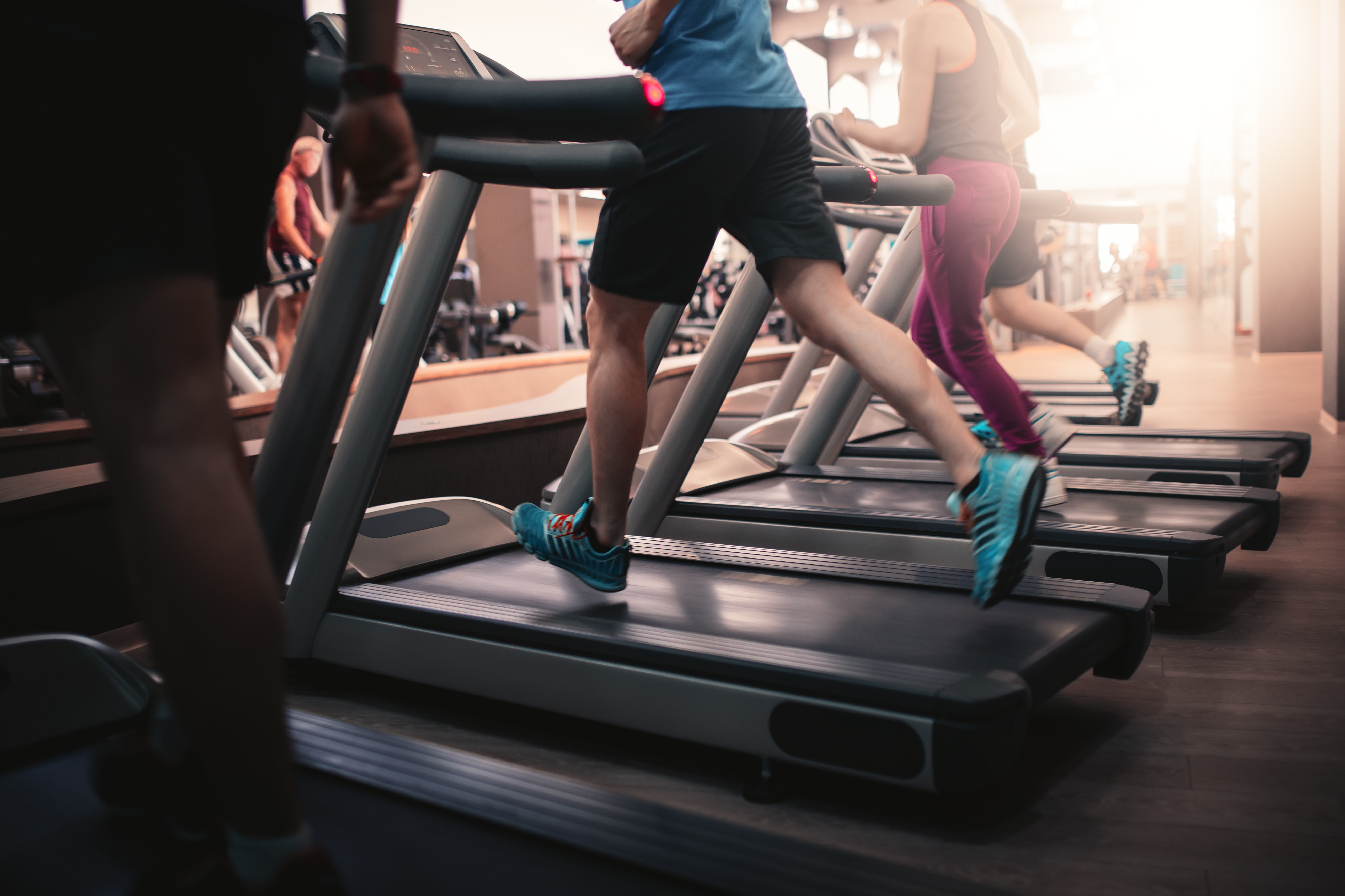 People running on treadmills in a modern gym, wearing athletic shoes and workout clothes, with exercise equipment visible in the background.
