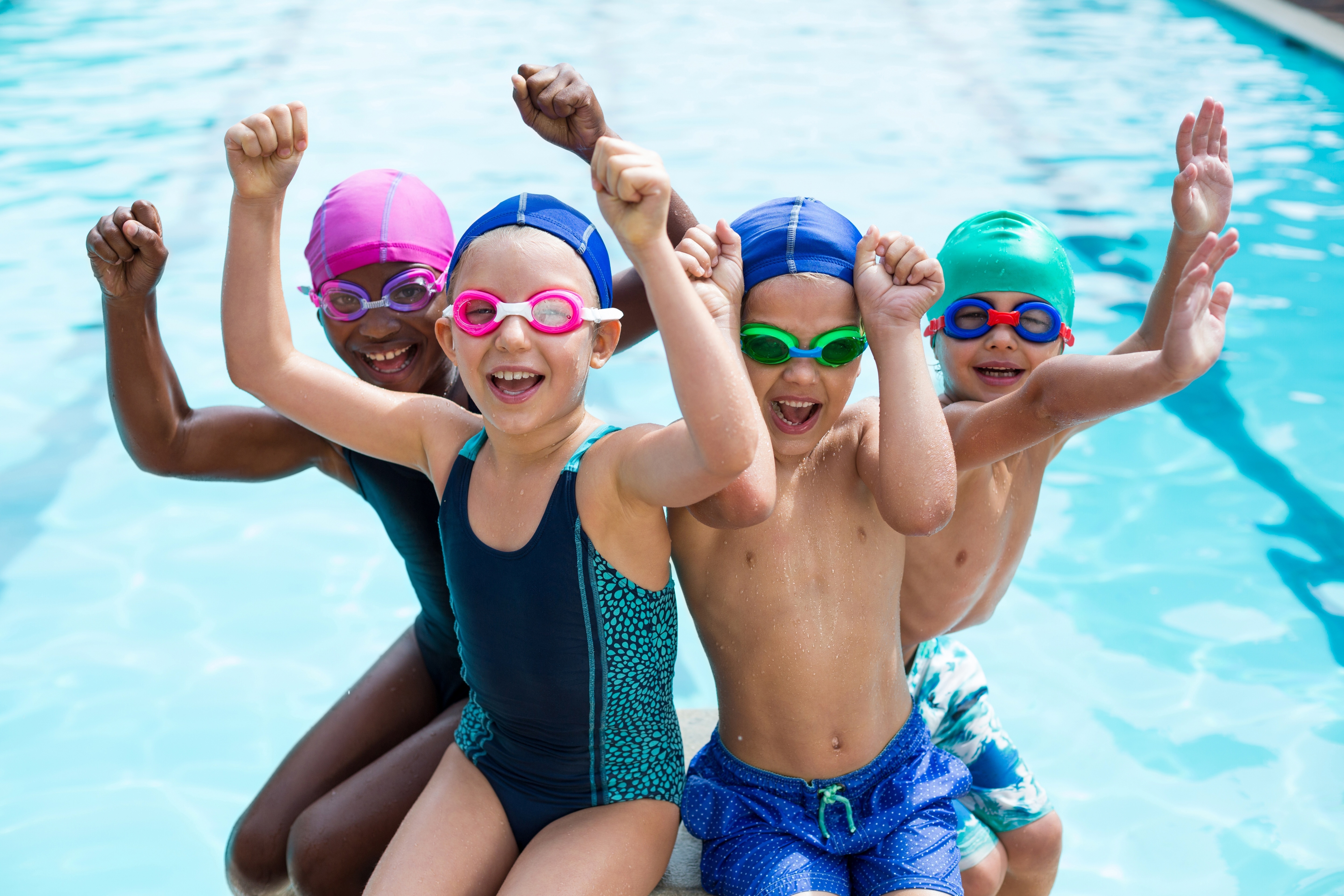A group of children wearing swim caps sit at the edge of a swimming pool with their arms raised in celebration, smiling and posing together during a swim session.