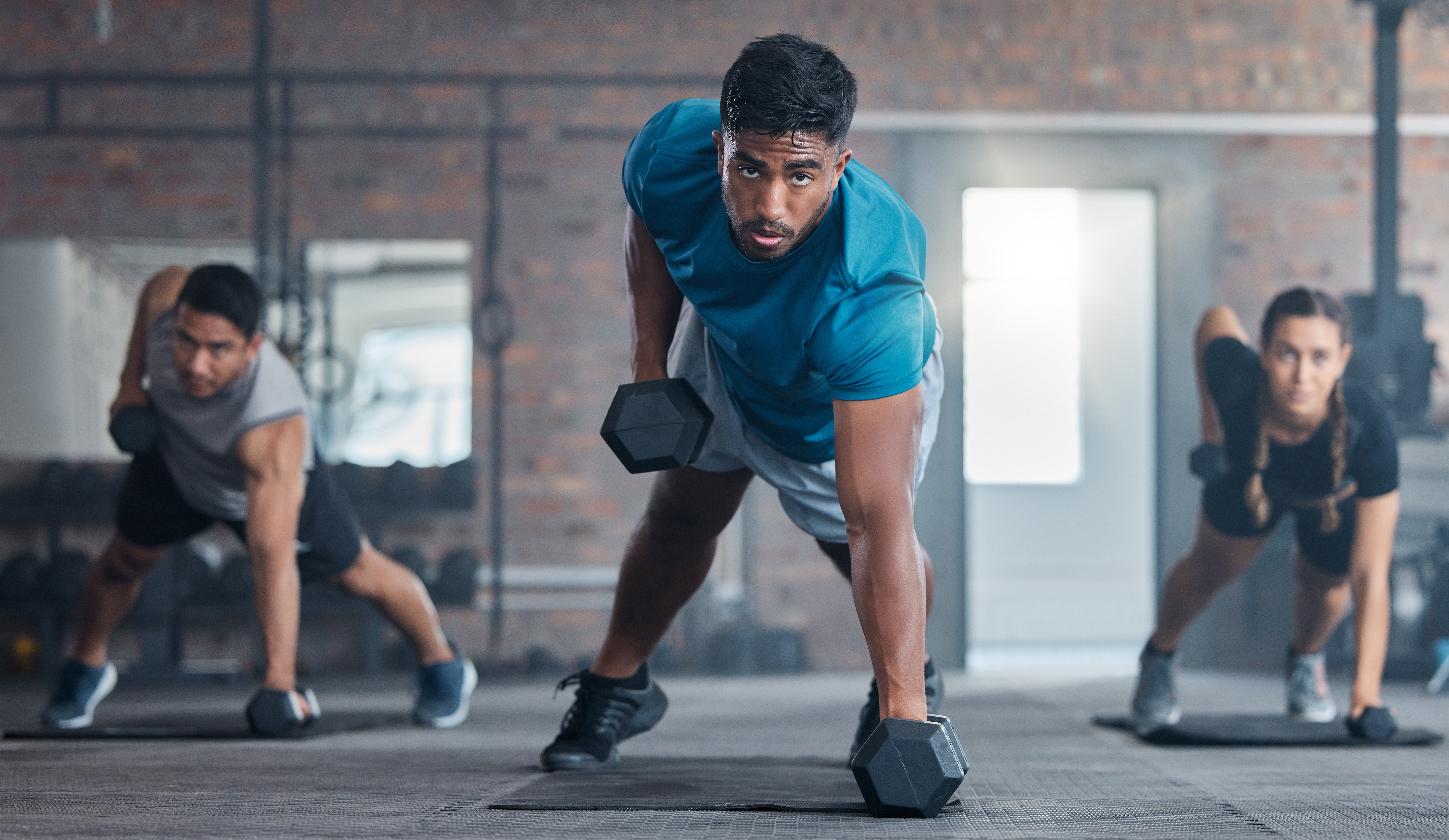 People in a gym performing a plank row exercise with dumbbells on a rubber floor.