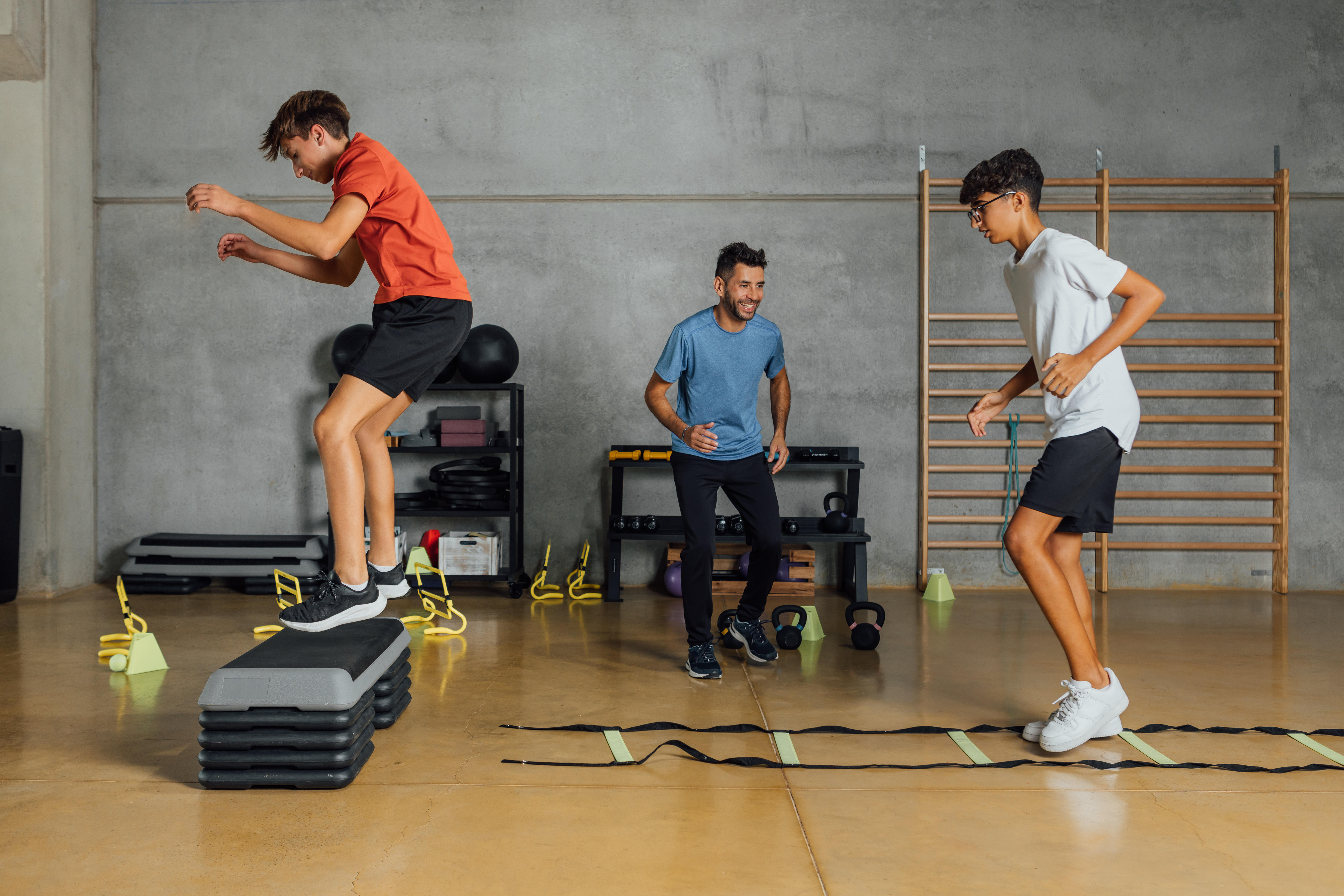 Two young people take part in a fitness session in a gym, one stepping onto stacked aerobic platforms and the other running through an agility ladder, while an instructor guides them.