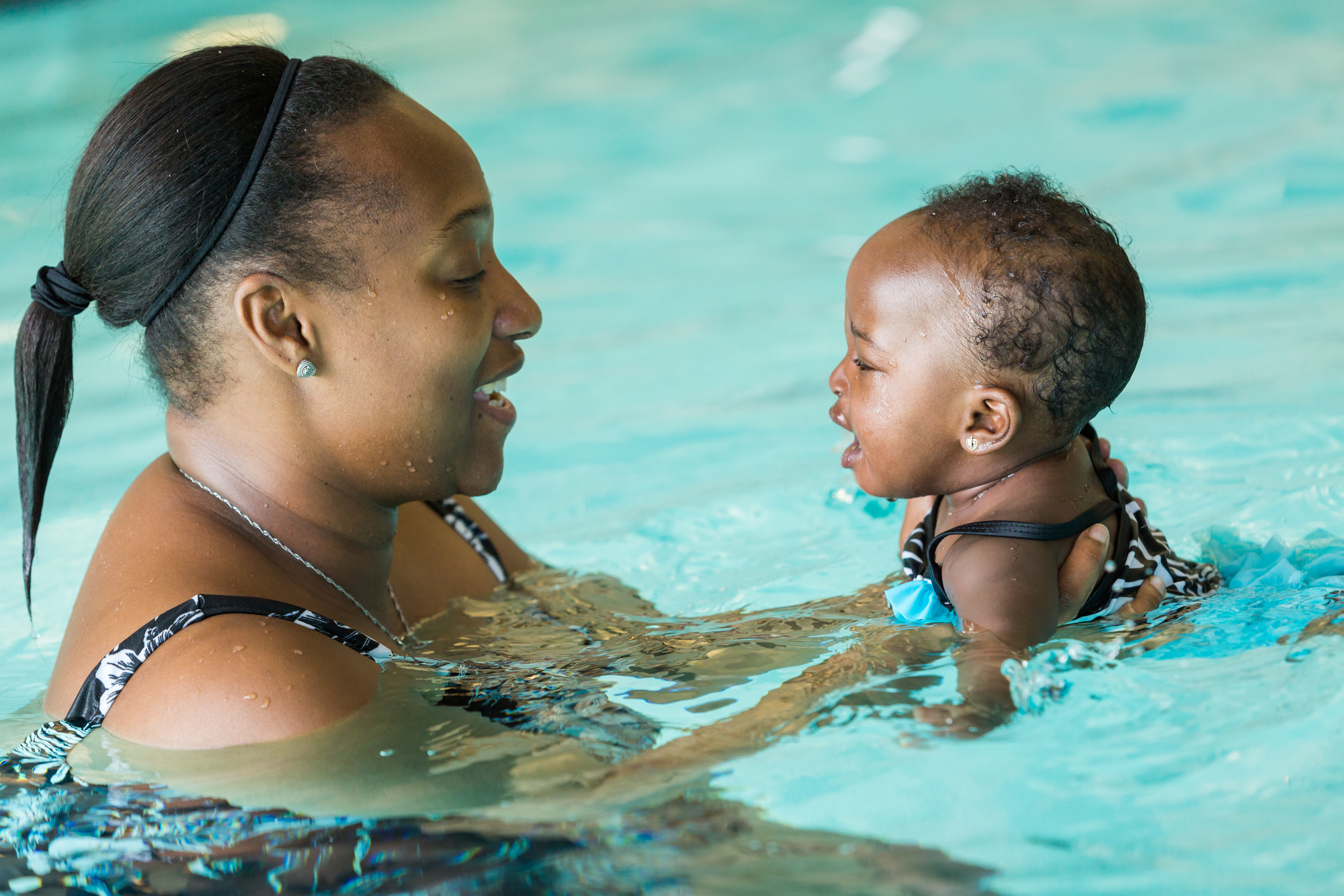 A caregiver in a swimming pool supports a small child as they face one another, with both partially immersed in the water.
