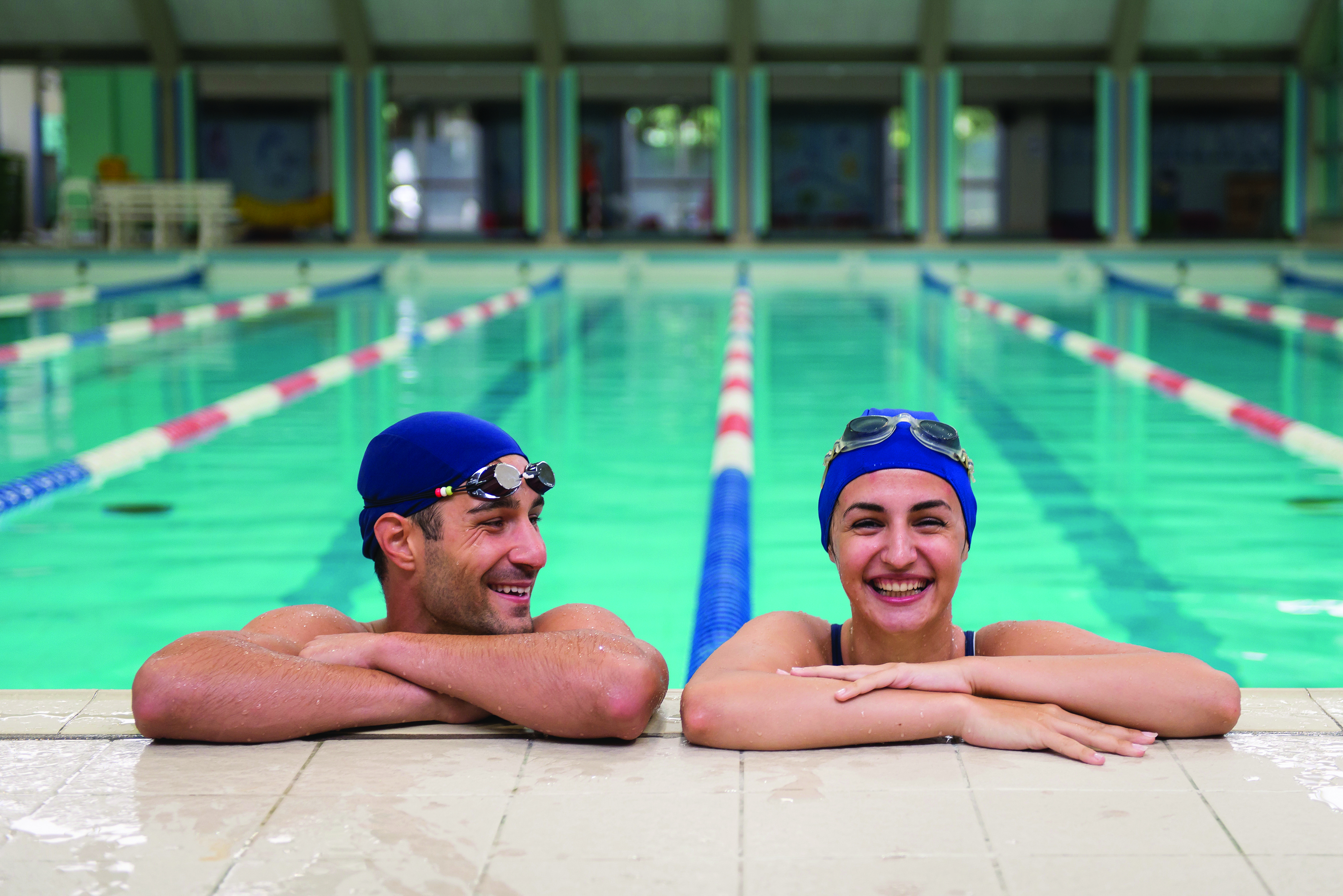 Two swimmers resting at the edge of an indoor swimming pool with lane dividers in the water.