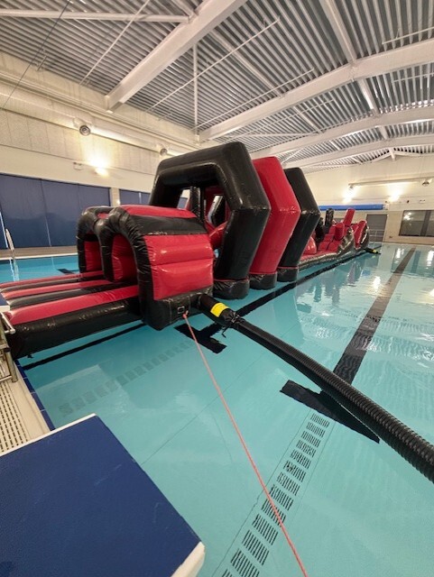 A large red‑and‑black inflatable obstacle course floats in an indoor swimming pool. The structure includes multiple arched sections and balance elements stretching across the width of the pool. The bright, modern pool hall has high ceilings with exposed beams and overhead lighting.