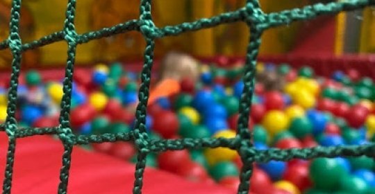 A close-up view of green netting in the foreground, with a colourful soft‑play ball pit and a child playing in the background out of focus.