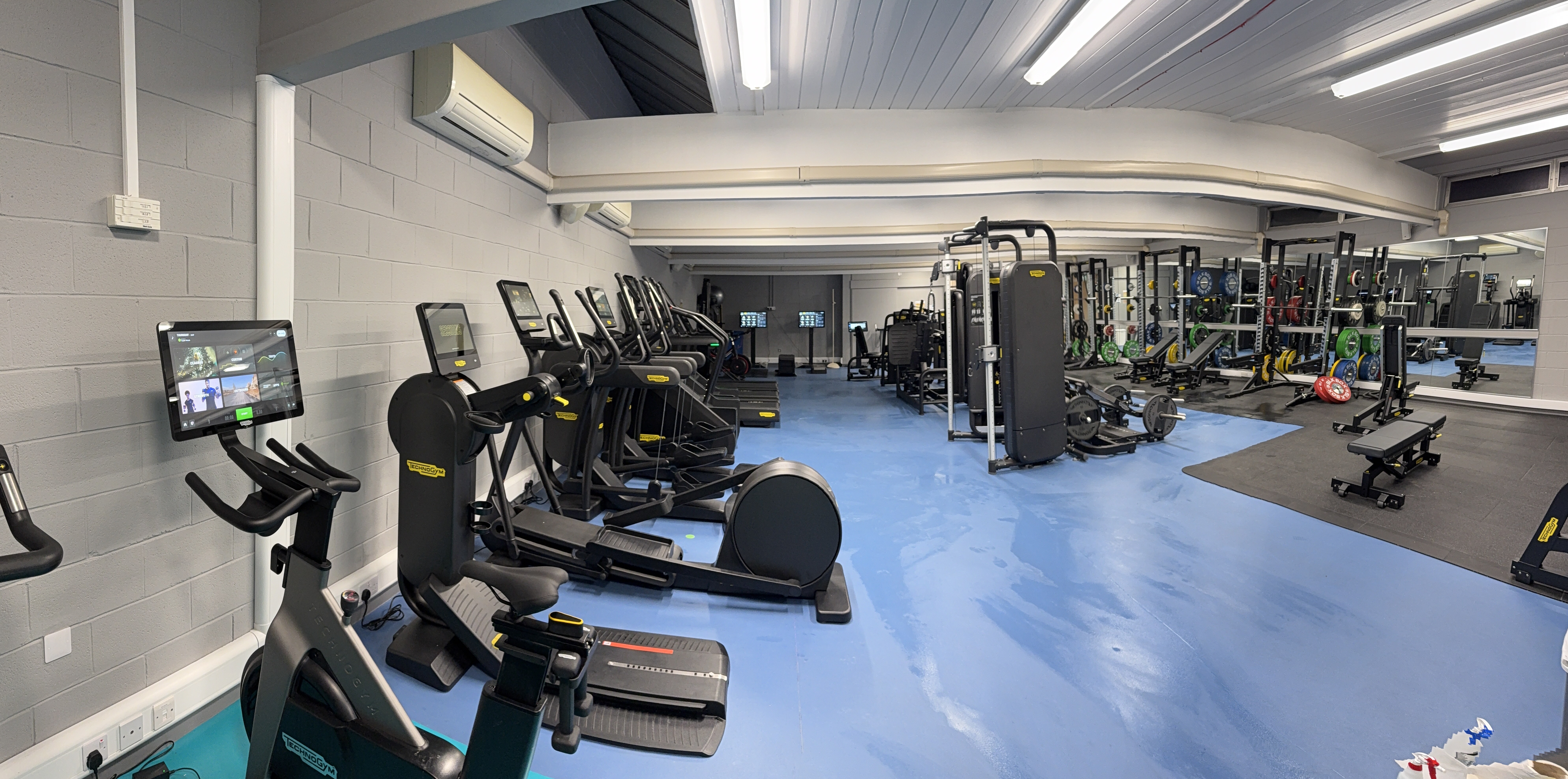 A spacious, modern gym with cardio machines lined up on the left, strength‑training equipment and free weights on the right, and bright overhead lighting illuminating the blue flooring and mirrored walls.