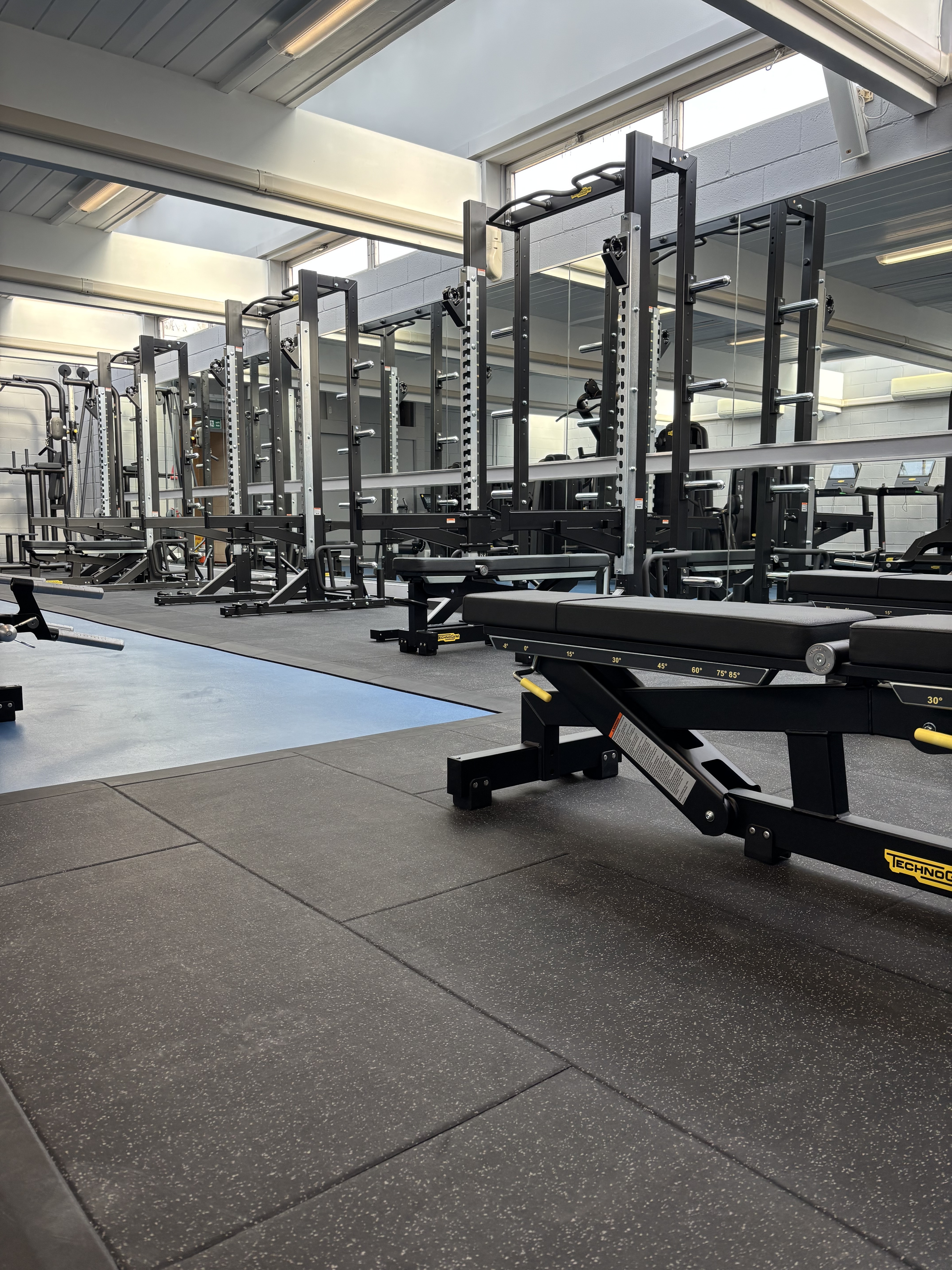 An adjustable weight bench sits on rubber flooring in a well‑lit gym area, with multiple squat racks, barbells, and mirrors visible in the background.