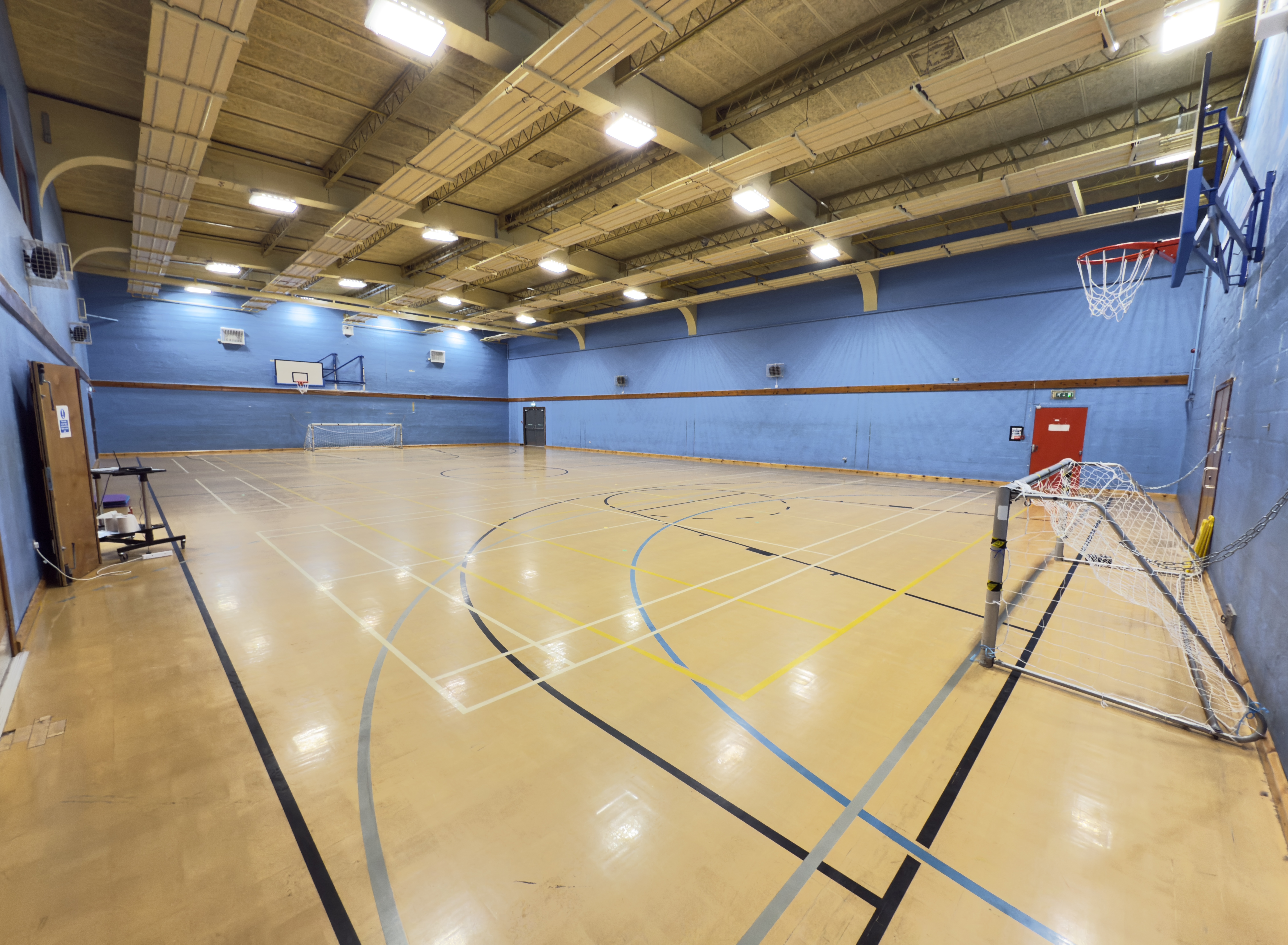 A large indoor sports hall with high ceilings, blue walls, and a polished floor marked for multiple sports, featuring basketball hoops and small goal nets at either end.