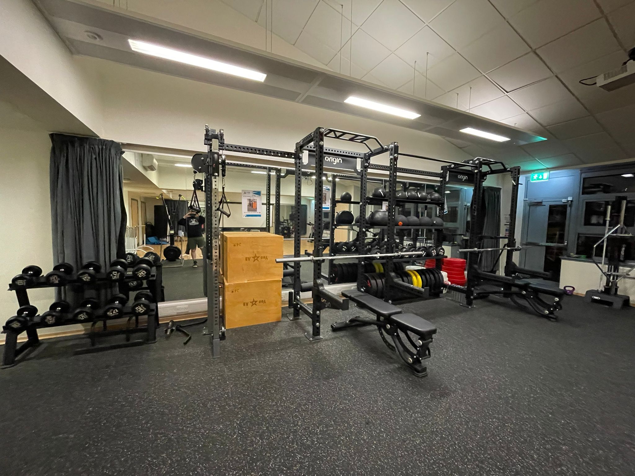 A well‑equipped gym studio featuring a large functional training rig with weight plates, kettlebells, and medicine balls. Dumbbells are neatly racked on the left, and adjustable benches are positioned in front of the rig on rubber flooring.