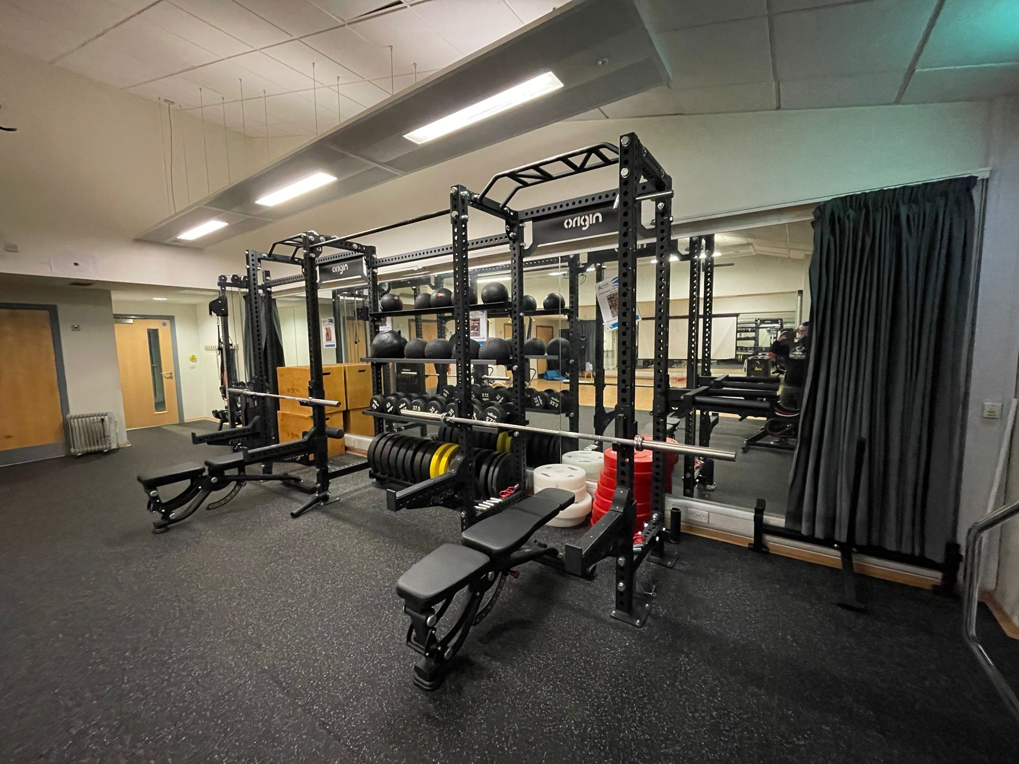 Strength‑training studio featuring a large Origin functional training rig with weight plates, kettlebells, medicine balls, and two adjustable benches on rubber flooring. A wall mirror and grey curtain sit behind the equipment under bright ceiling lights.