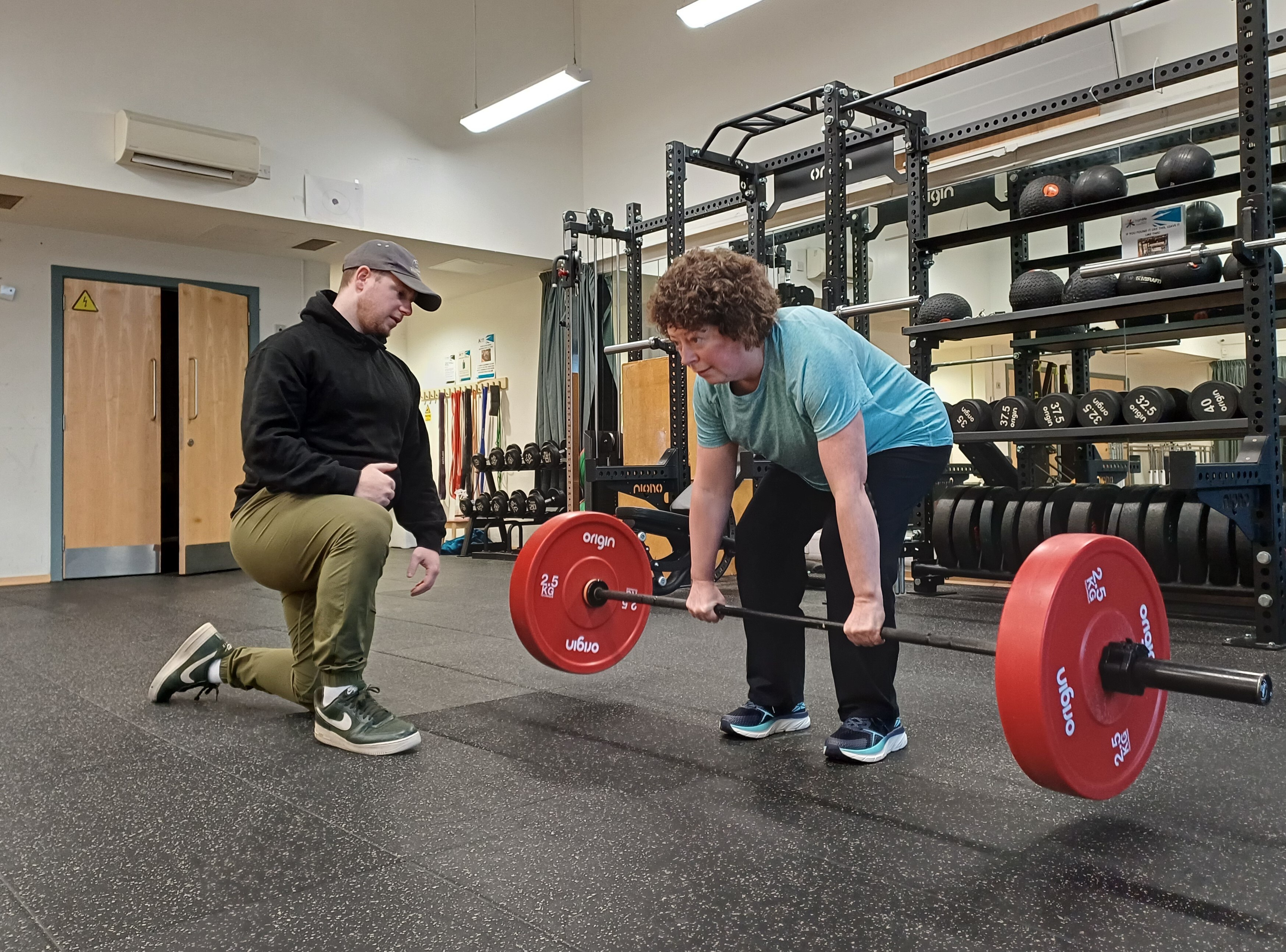 A person performs a deadlift with a barbell loaded with red weight plates while an instructor kneels nearby providing guidance. The exercise takes place in a gym studio with a functional training rig, weight plates, and kettlebells in the background.