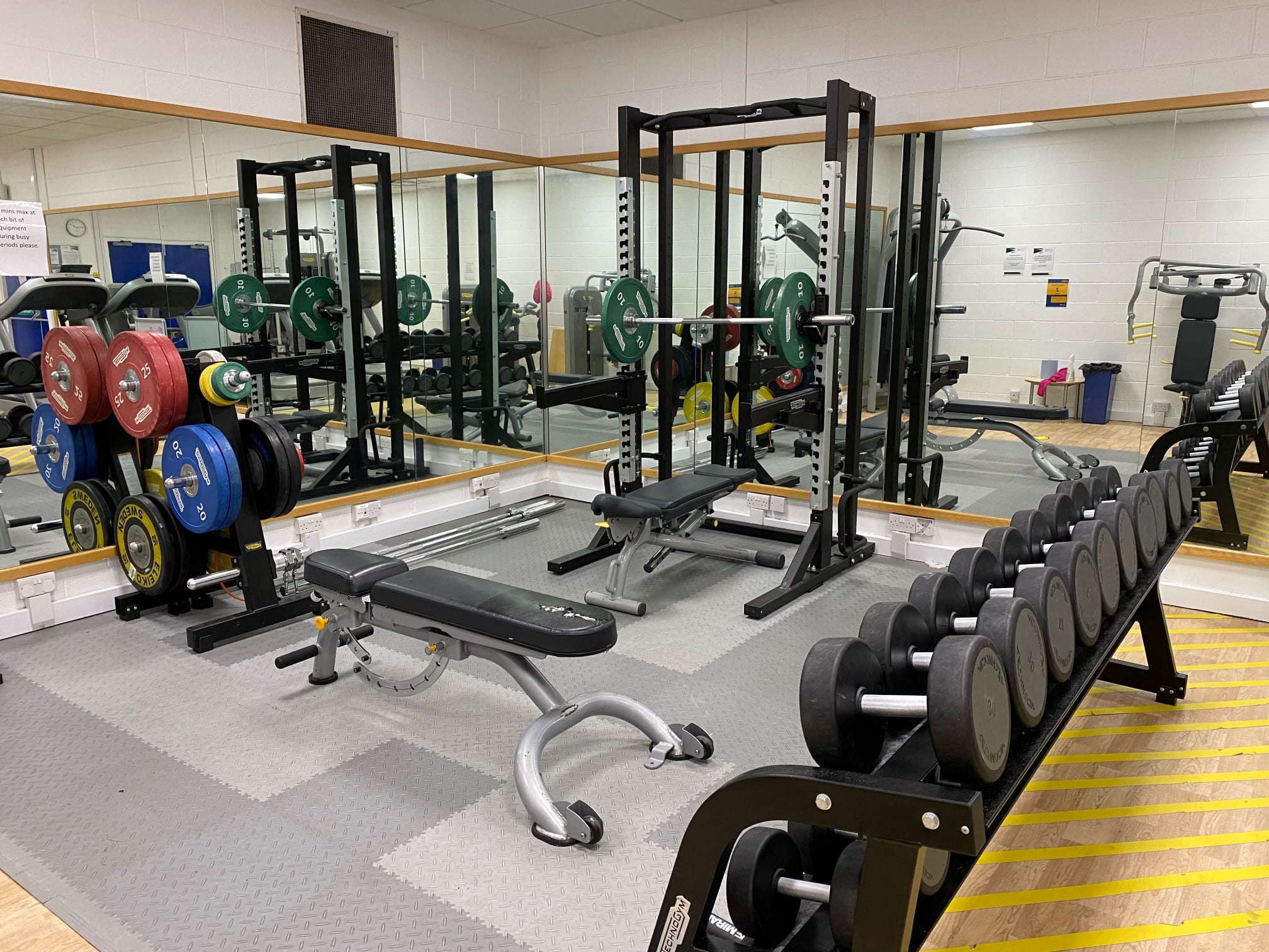 Gym weightlifting area with barbells, benches, and wall mirrors.