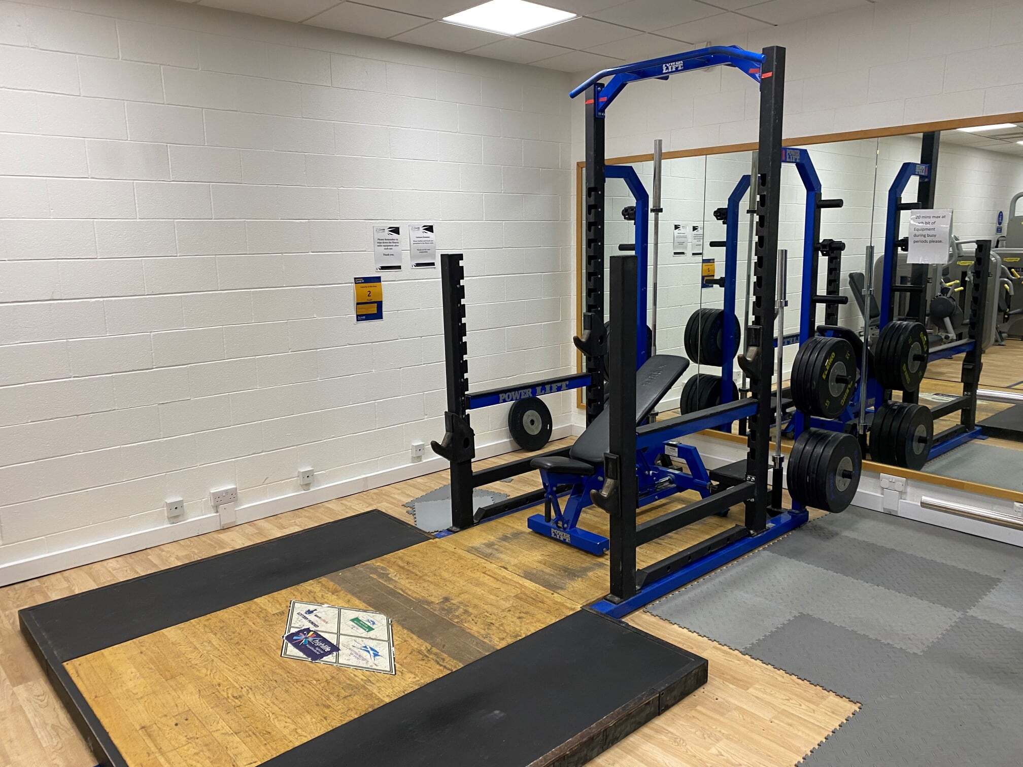 A weightlifting area featuring a blue power rack with an adjustable bench, loaded weight plates, and a wooden lifting platform. The setup is positioned next to a large wall mirror in a bright gym room with white brick walls and light flooring.