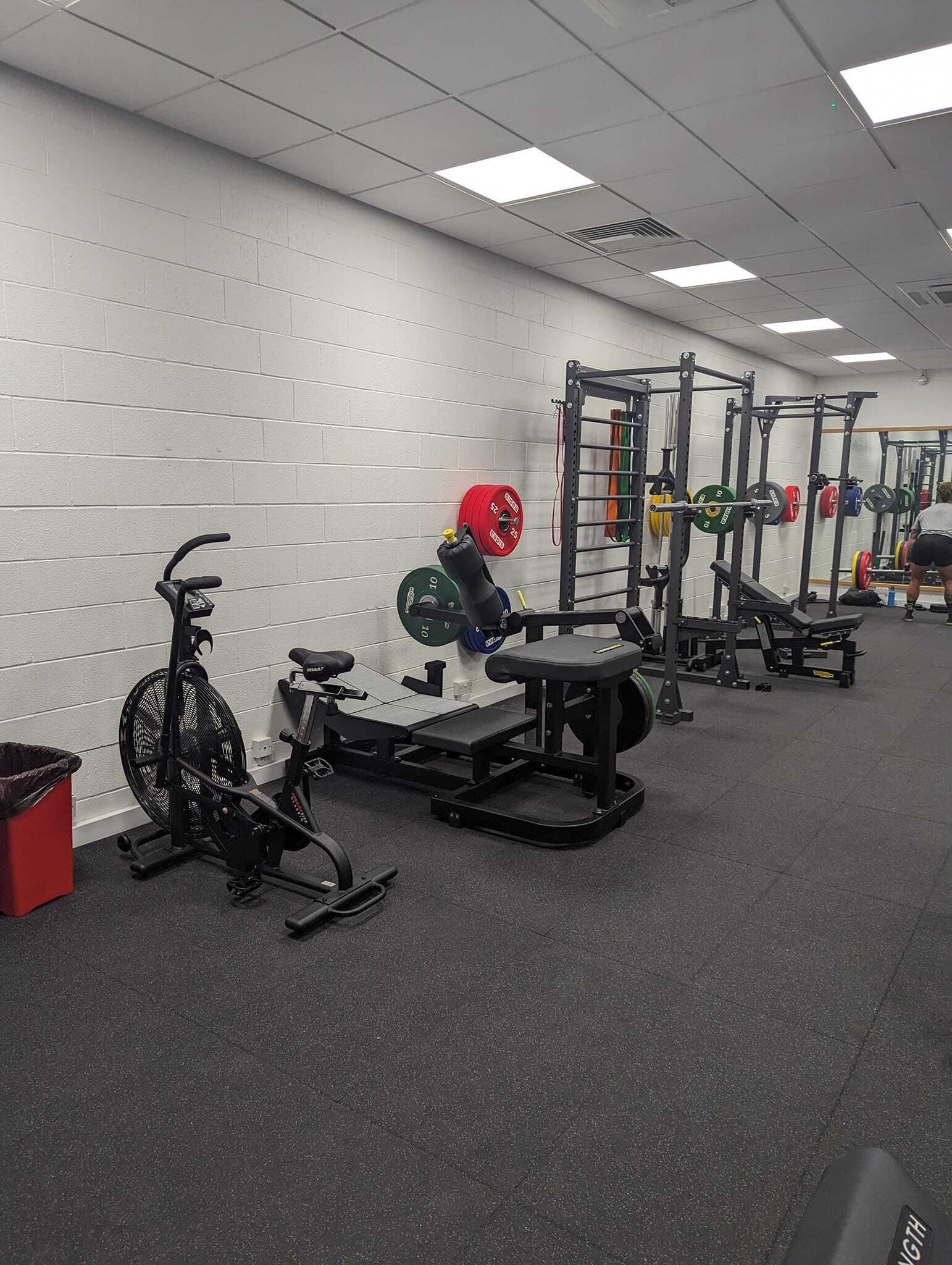 Strength‑training area with an assault bike, weight benches, hip thrust machine, and multiple squat racks equipped with colourful weight plates. The space features rubber flooring, white brick walls, and bright overhead lighting.