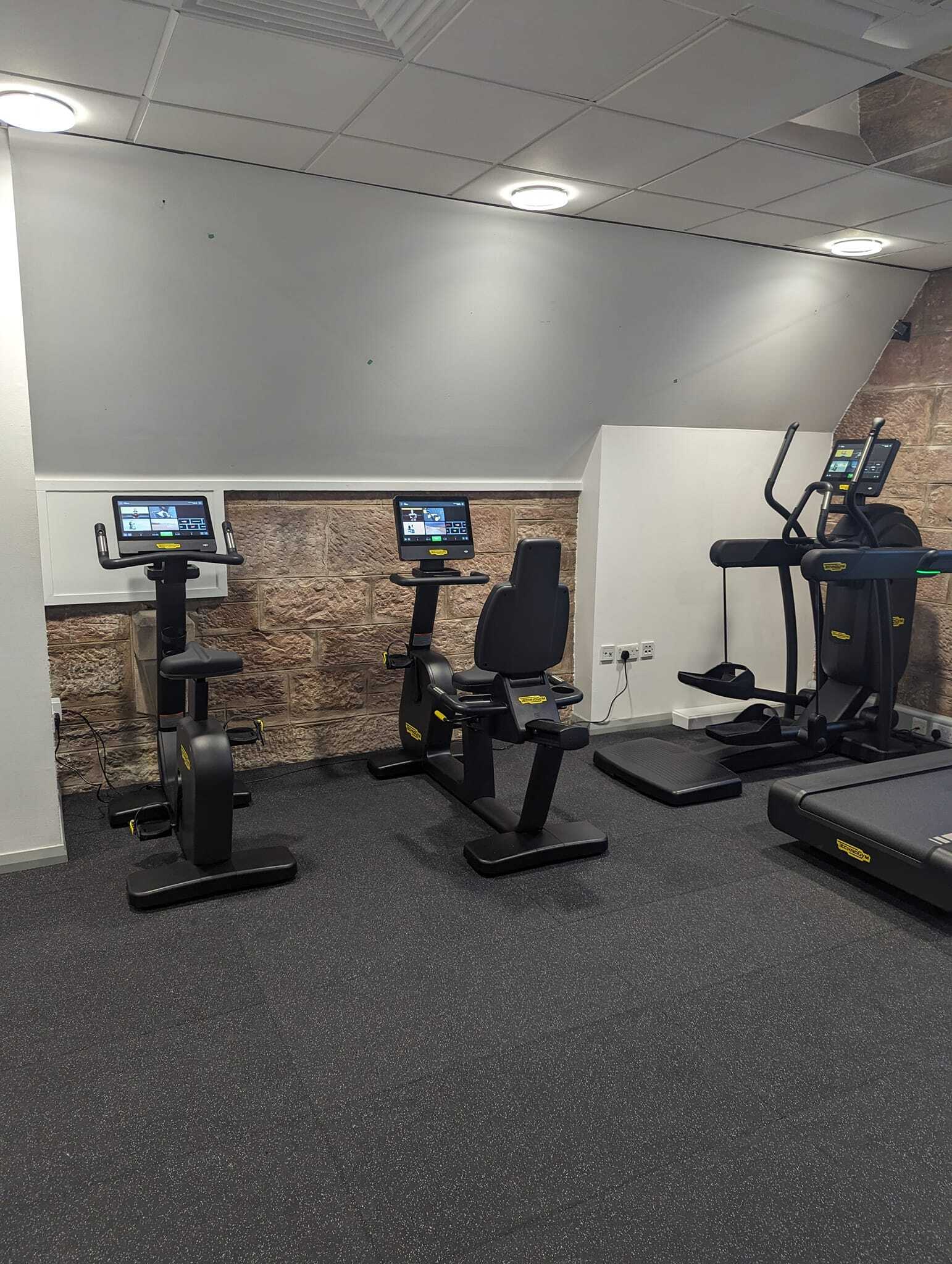 Cardio area featuring two upright exercise bikes, a recumbent bike, and a cross‑trainer, all equipped with digital screens. The equipment is arranged against a wall with stone detailing under bright ceiling lights on dark rubber flooring.