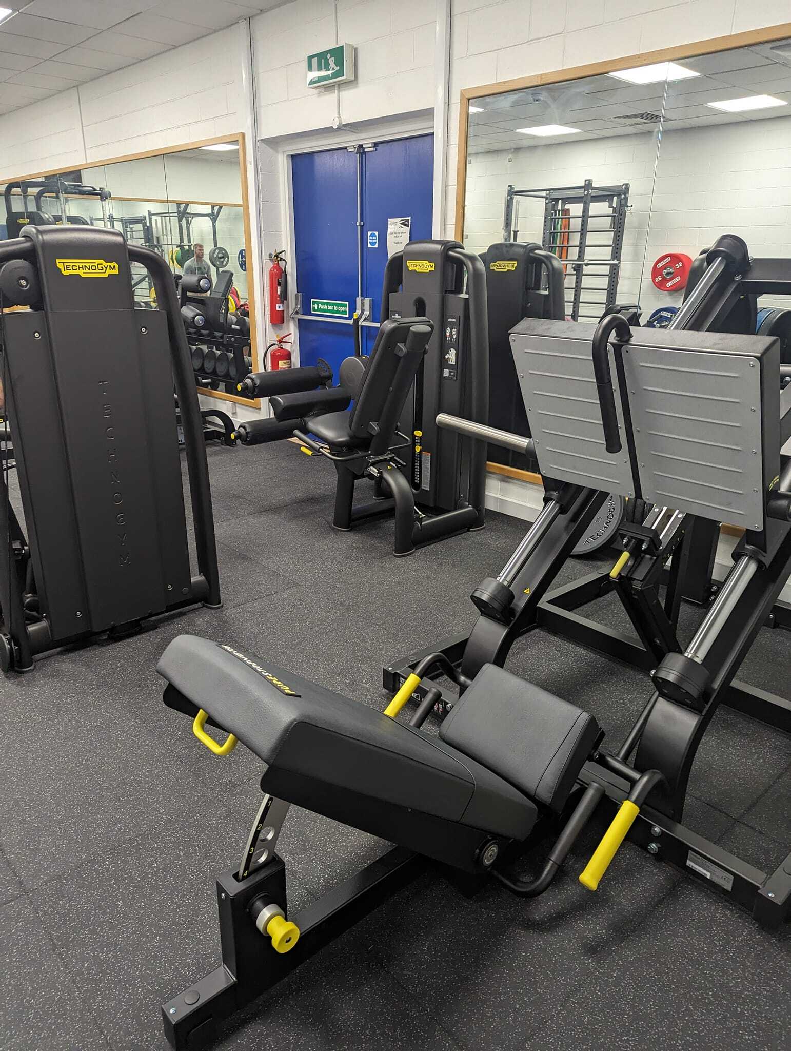 Gym area with several Technogym strength machines, including a leg press, leg curl, and adjustable bench, arranged on rubber flooring. Mirrors line the wall behind the equipment,