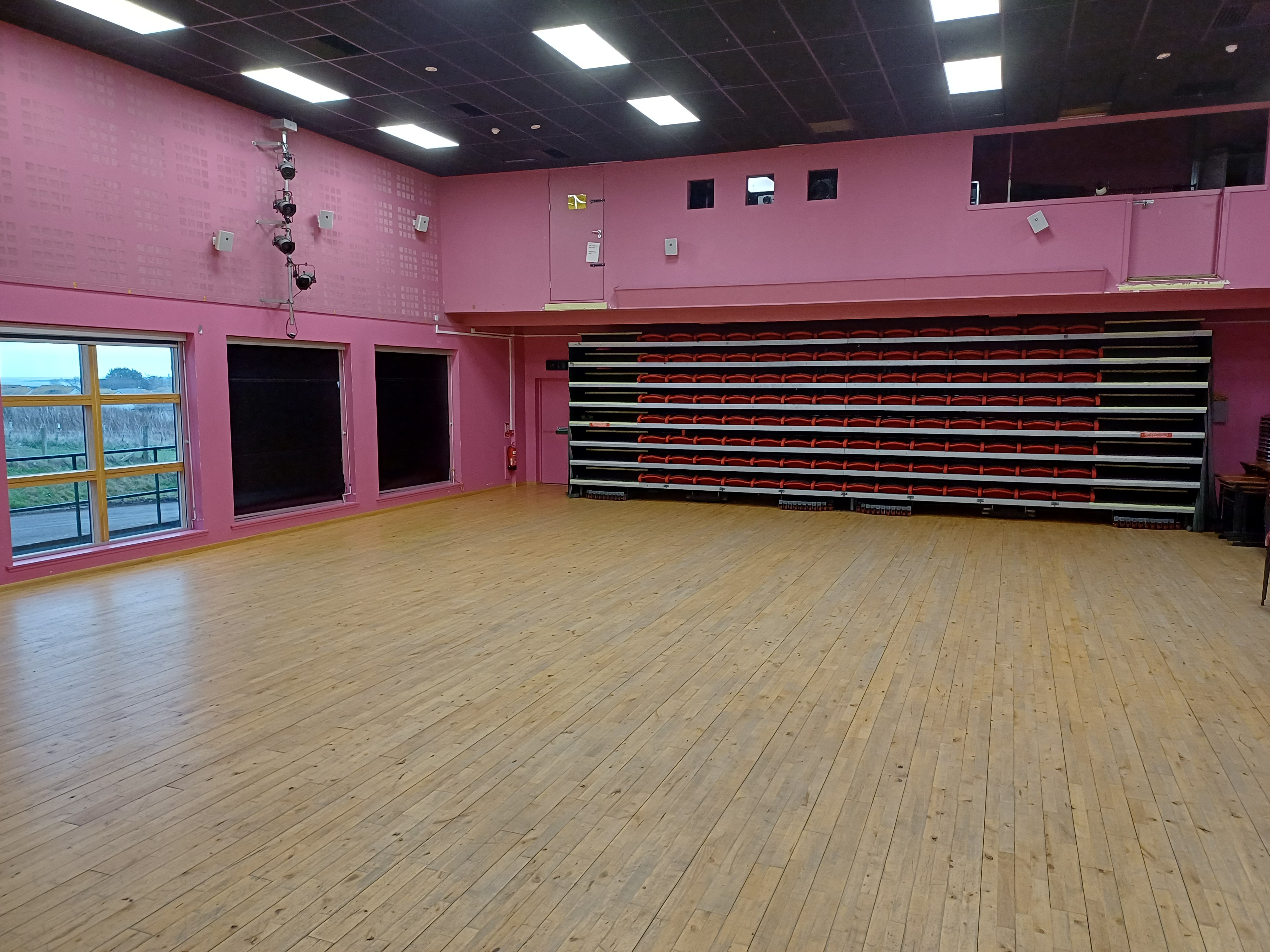 A large empty hall with pink walls, a black ceiling, and a wooden floor. Along the back wall is a folded stack of tiered seating with red and black seats.