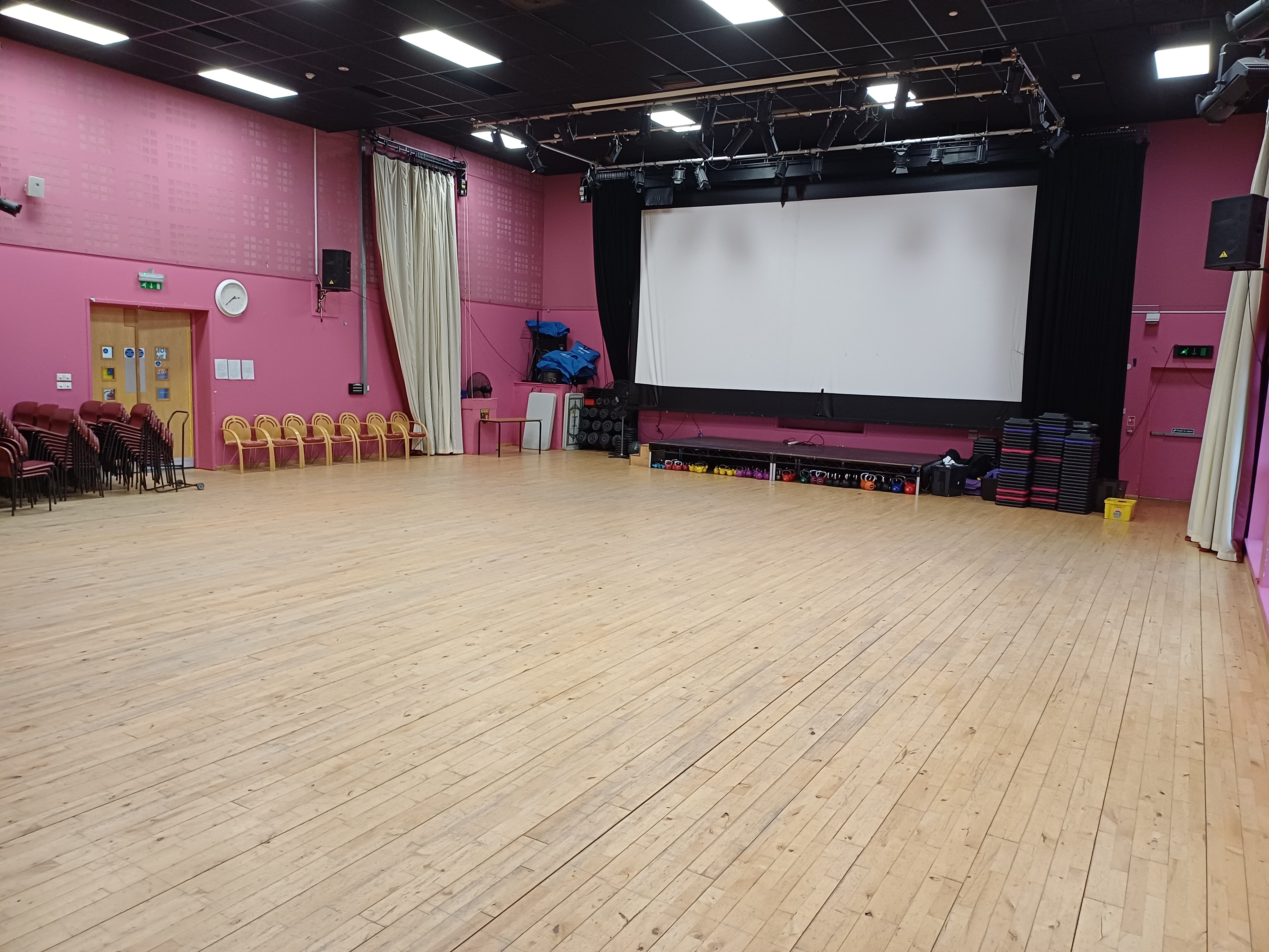A large hall with pink walls, a wooden floor, and a black ceiling with bright rectangular lights. At the far end is a stage area featuring a large pull-down projection screen framed by black curtains; the screen appears blank and ready for use.