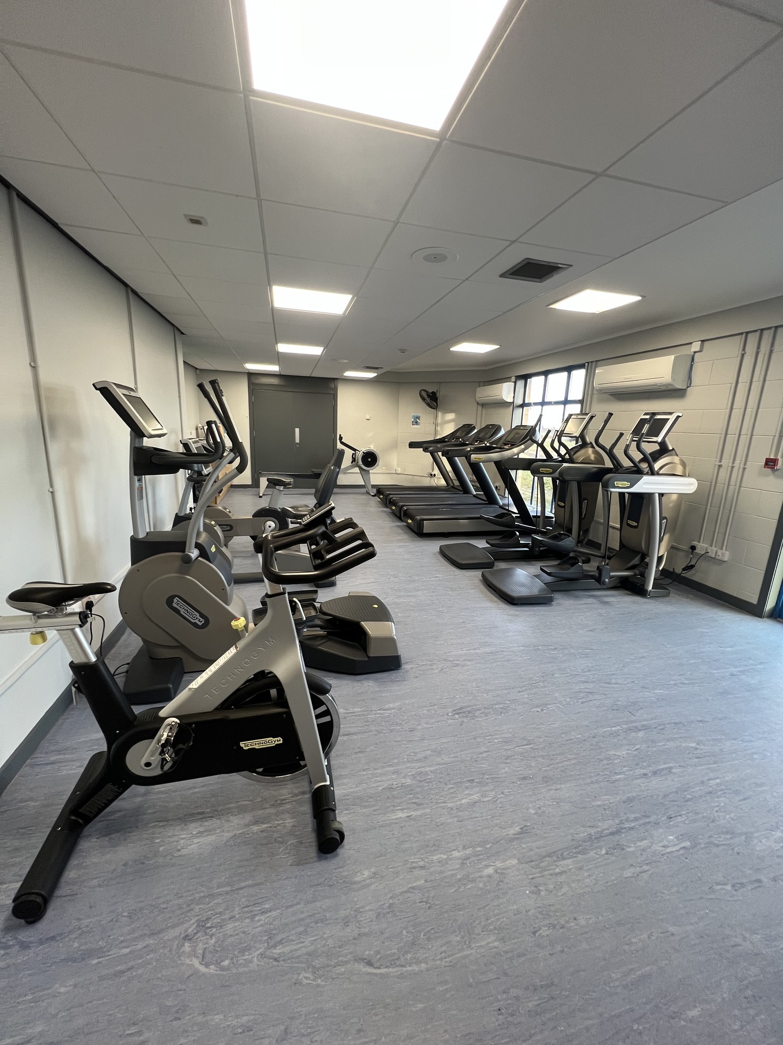 A bright, modern gym room with several pieces of cardio equipment arranged in rows, including exercise bikes, rowing machines, and treadmills. The space has a clean blue floor, white ceiling panels with fluorescent lighting, and a large window along one wall.