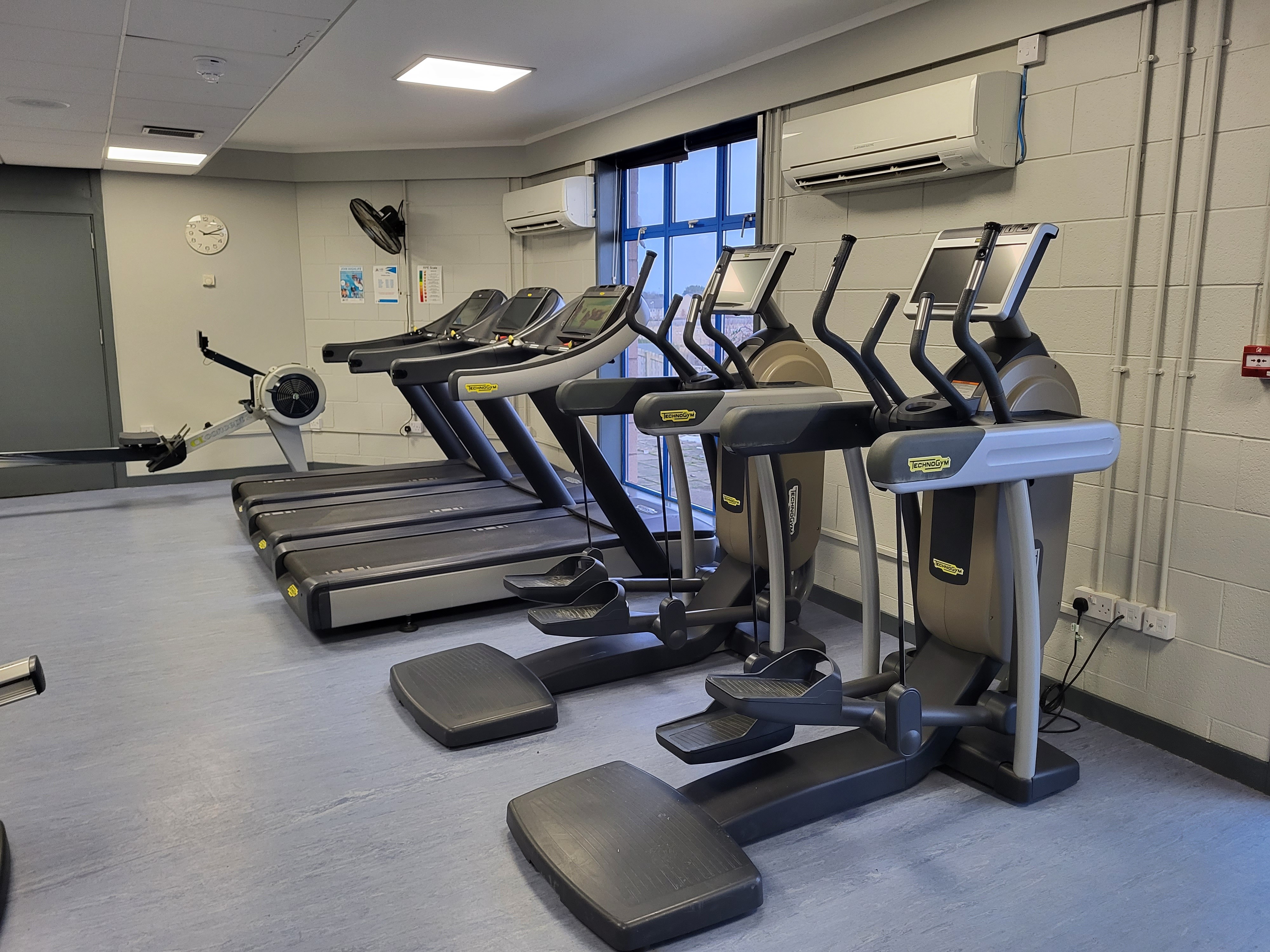 A gym cardio area featuring several treadmills and elliptical machines arranged in rows beside a large window. The room has light-colored flooring, wall‑mounted air conditioning units, and bright overhead lighting.