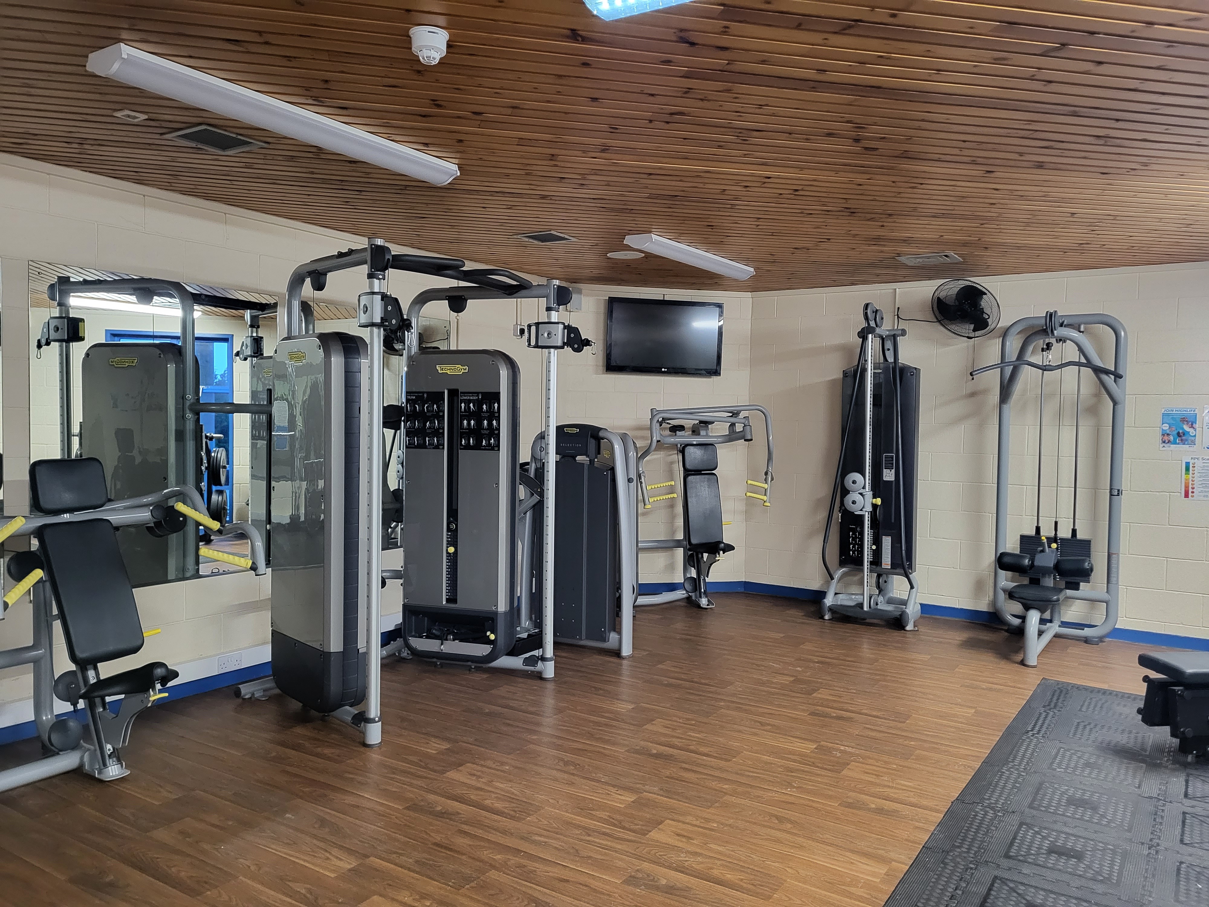 A gym room with several strength‑training machines, including cable machines, a seated chest press, and other resistance equipment arranged along the walls. The space features wood‑effect flooring, mirrors, and a wooden ceiling with overhead lighting.