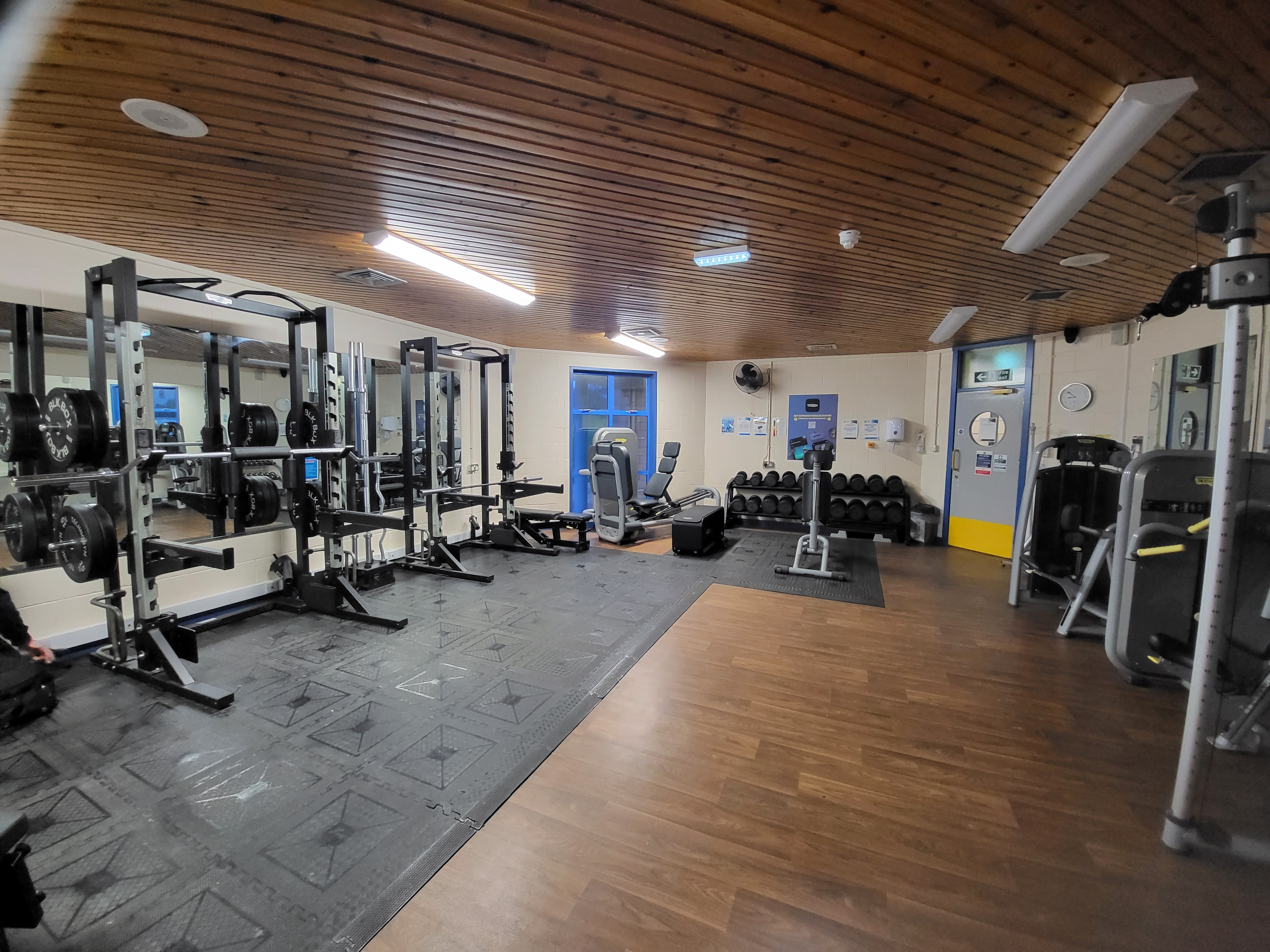 A strength training room with weight racks, barbells, dumbbells, and various gym machines arranged around the space. The room has a wooden ceiling, a mix of rubber and wood flooring, large mirrors on one wall, and a window providing natural light.