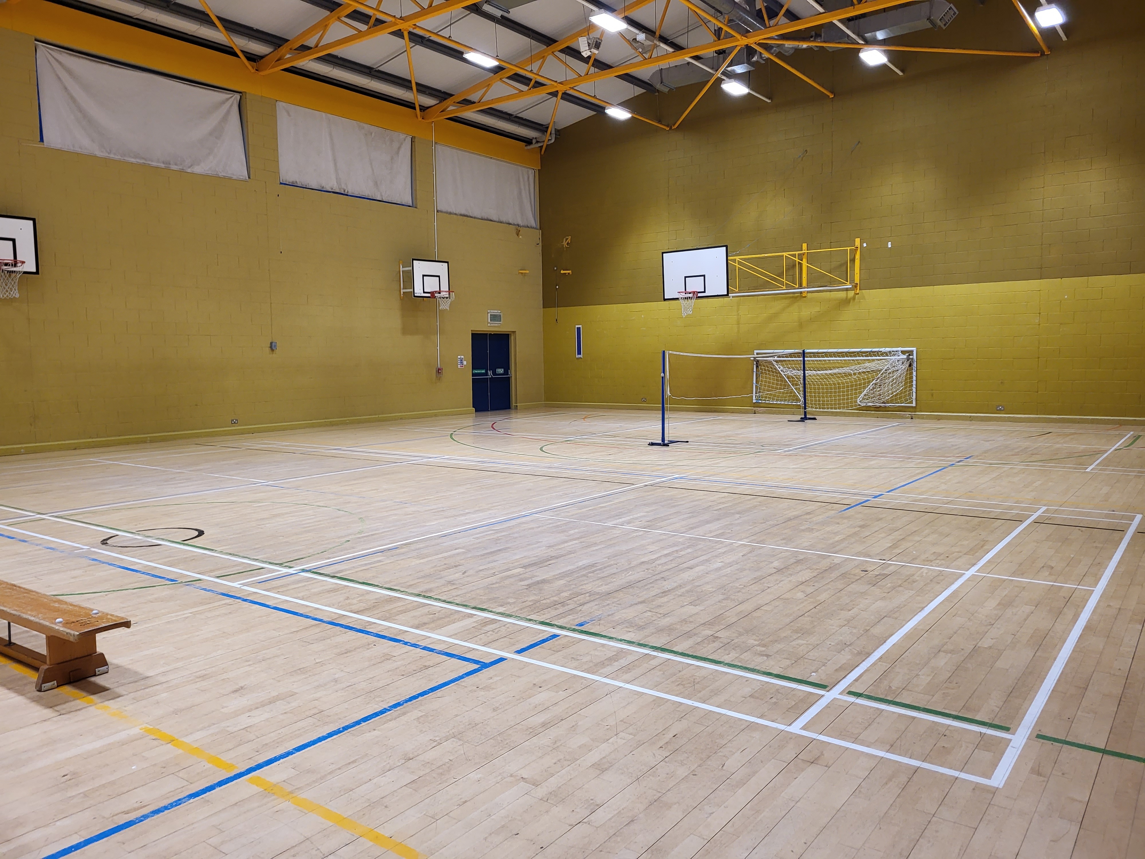 Indoor sports hall with a wooden floor, marked courts, tennis nets set up across the space, two small football goals, and basketball hoops mounted on the walls under a high yellow‑beamed ceiling.