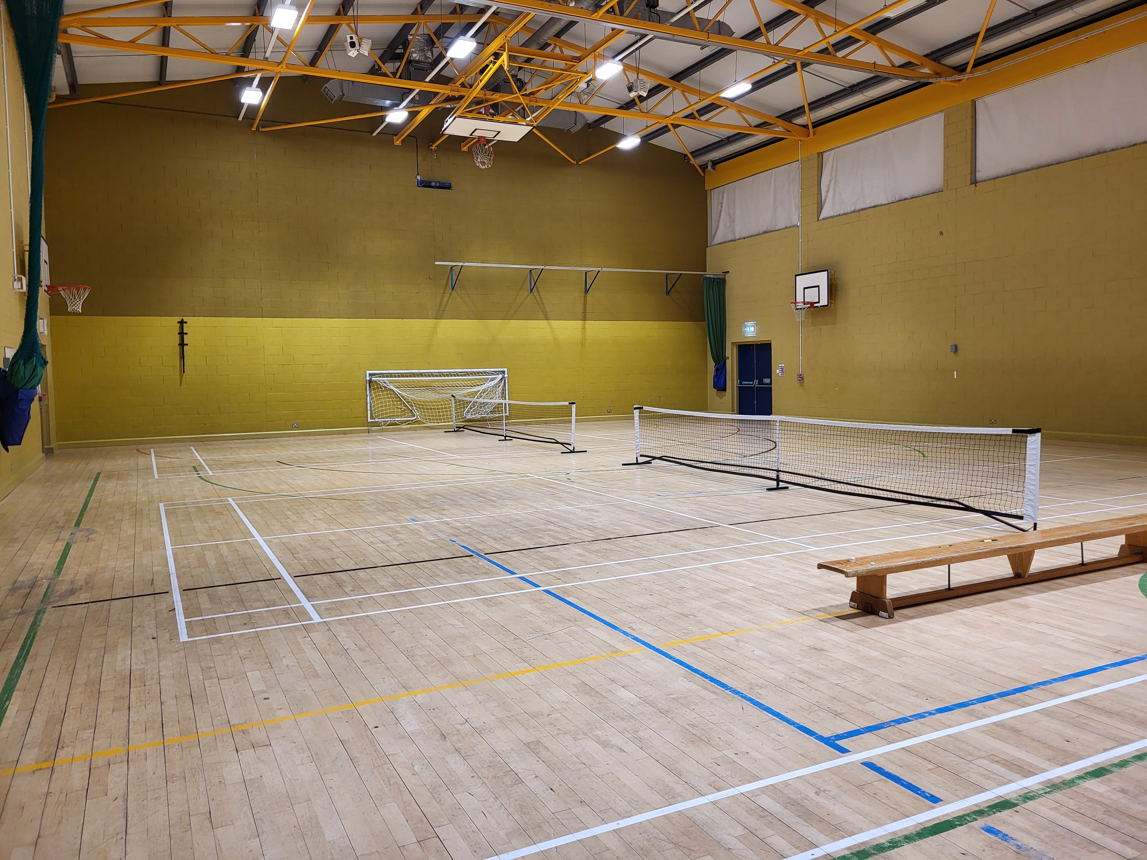 Indoor sports hall with a wooden floor, marked courts, tennis nets set up across the space, two small football goals, and basketball hoops mounted on the walls under a high yellow‑beamed ceiling.
