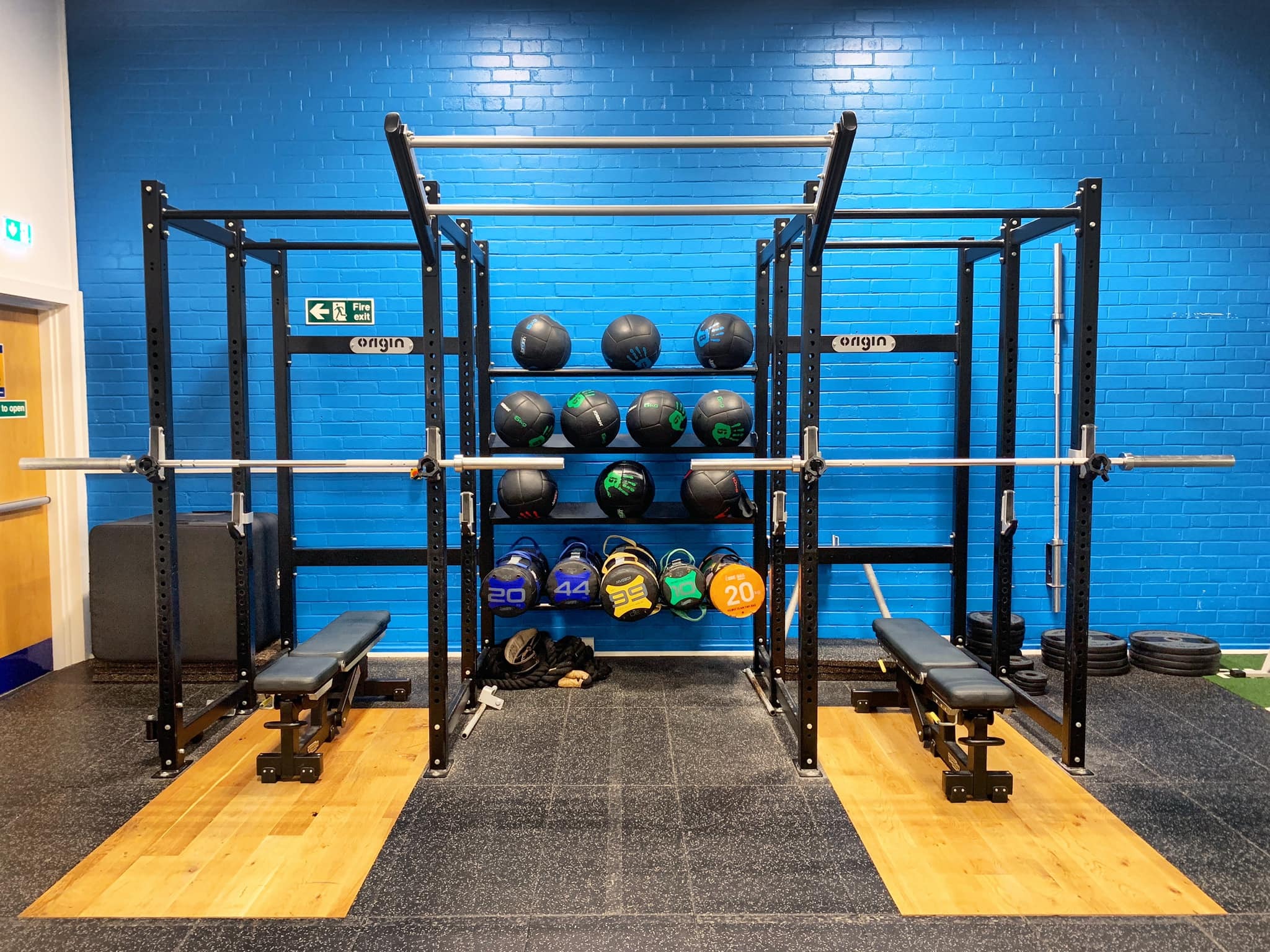 A gym setup featuring a black metal power rack with two weight benches on wooden platforms. Behind the rack, shelves hold medicine balls and coloured weighted bags. A bright blue brick wall forms the backdrop, and barbells are racked across the frame.