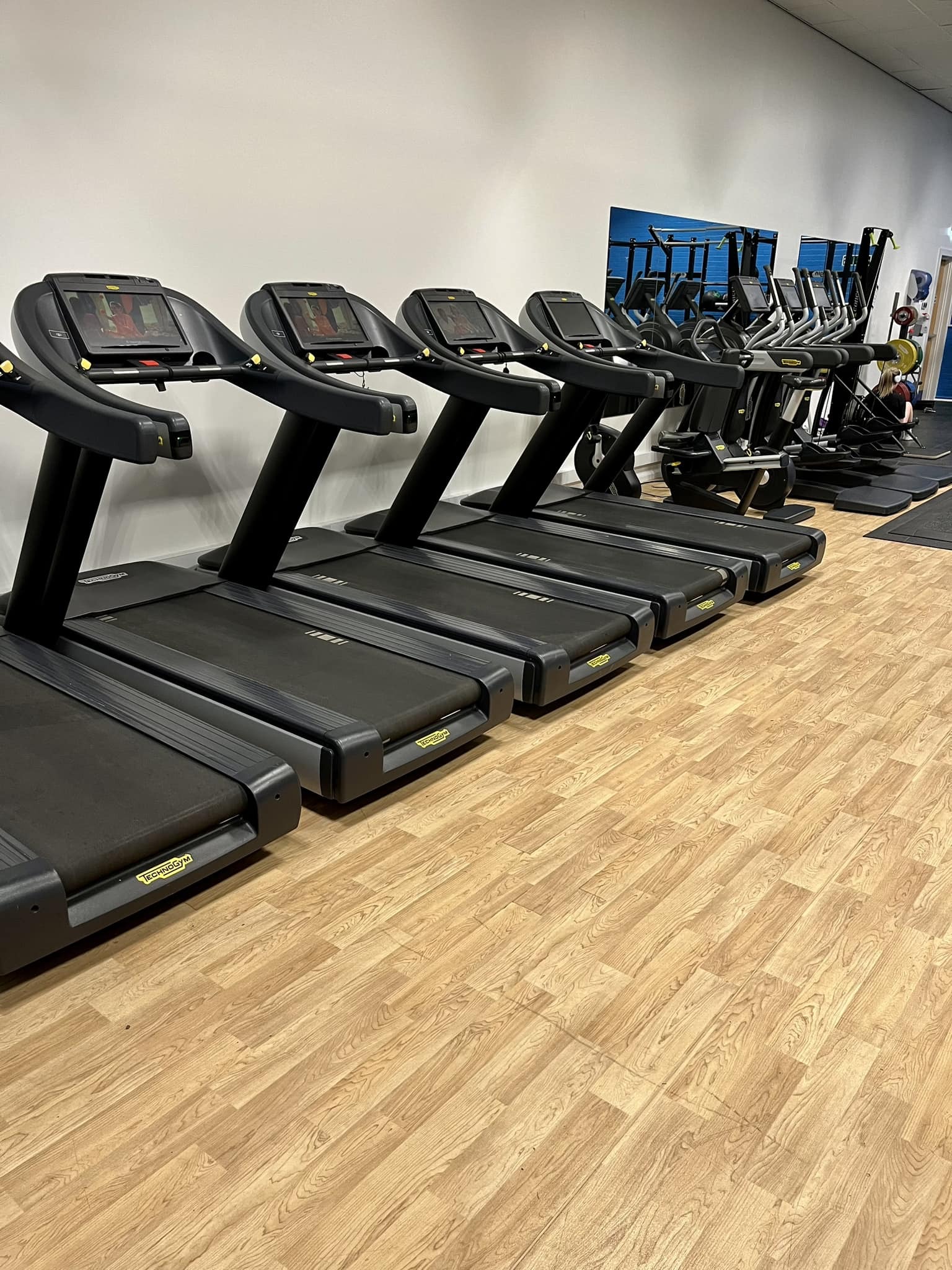 A row of black treadmills lined up along a white wall in a gym, with wooden flooring beneath. Behind the treadmills are several elliptical machines and other cardio equipment.