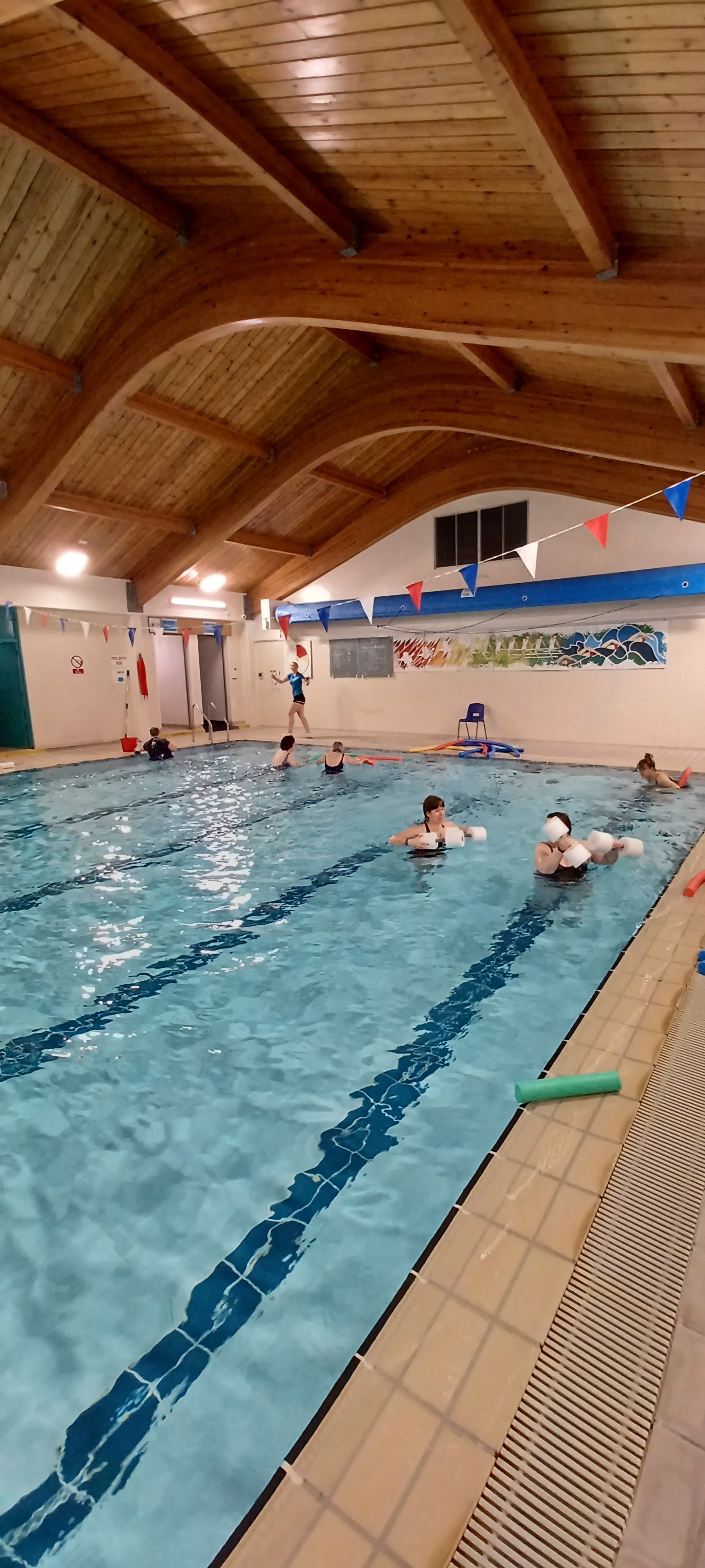 An indoor swimming pool with lane markings, where several people are taking part in a swim session using floats and pool noodles. The pool is surrounded by tiled flooring, and the space has a high, curved wooden ceiling with overhead lighting. Colourful bunting hangs across the pool area, and a mural is visible on the far wall.