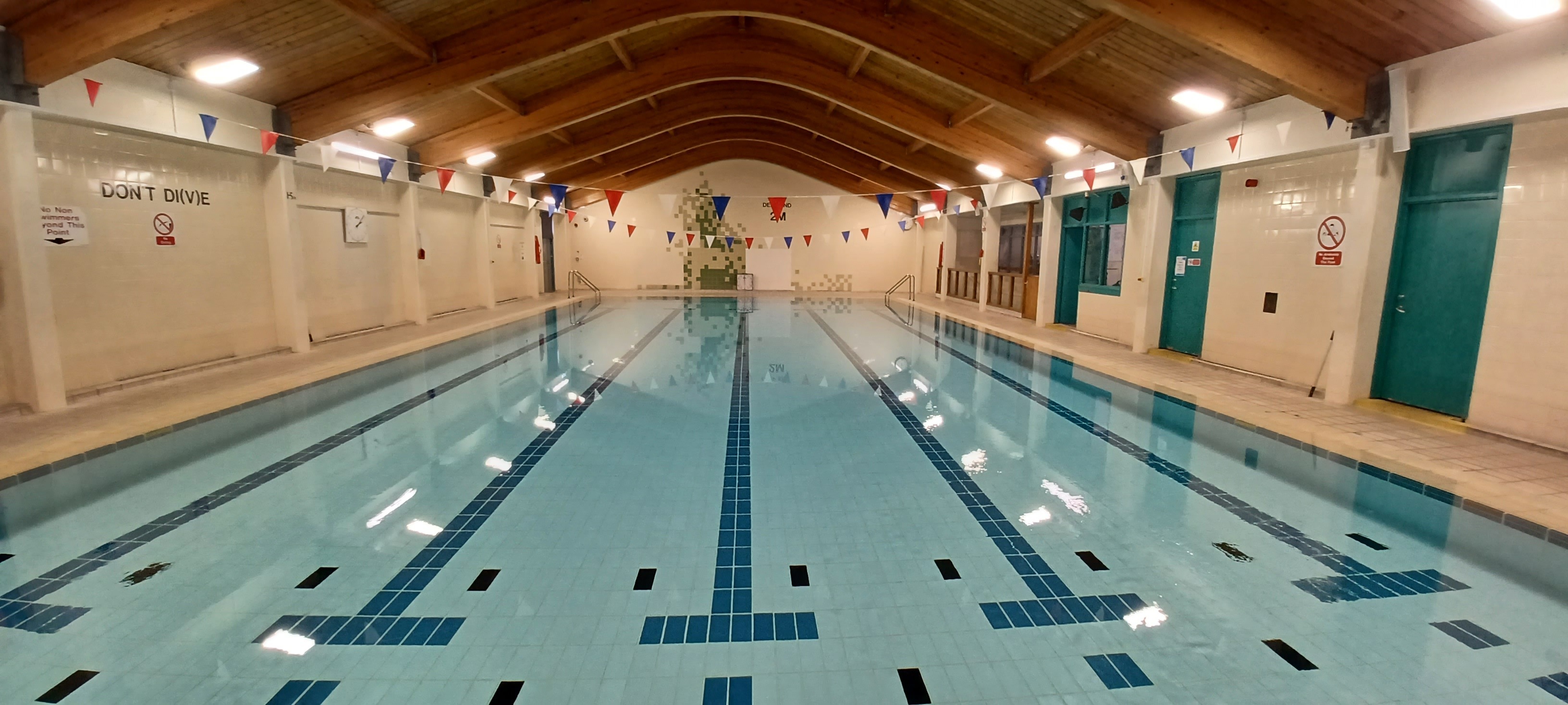 An indoor swimming pool with clear blue water and lane markings, viewed from one end looking straight down the length of the pool. The pool is surrounded by cream‑coloured tiles and has a high arched wooden ceiling with bright overhead lighting.