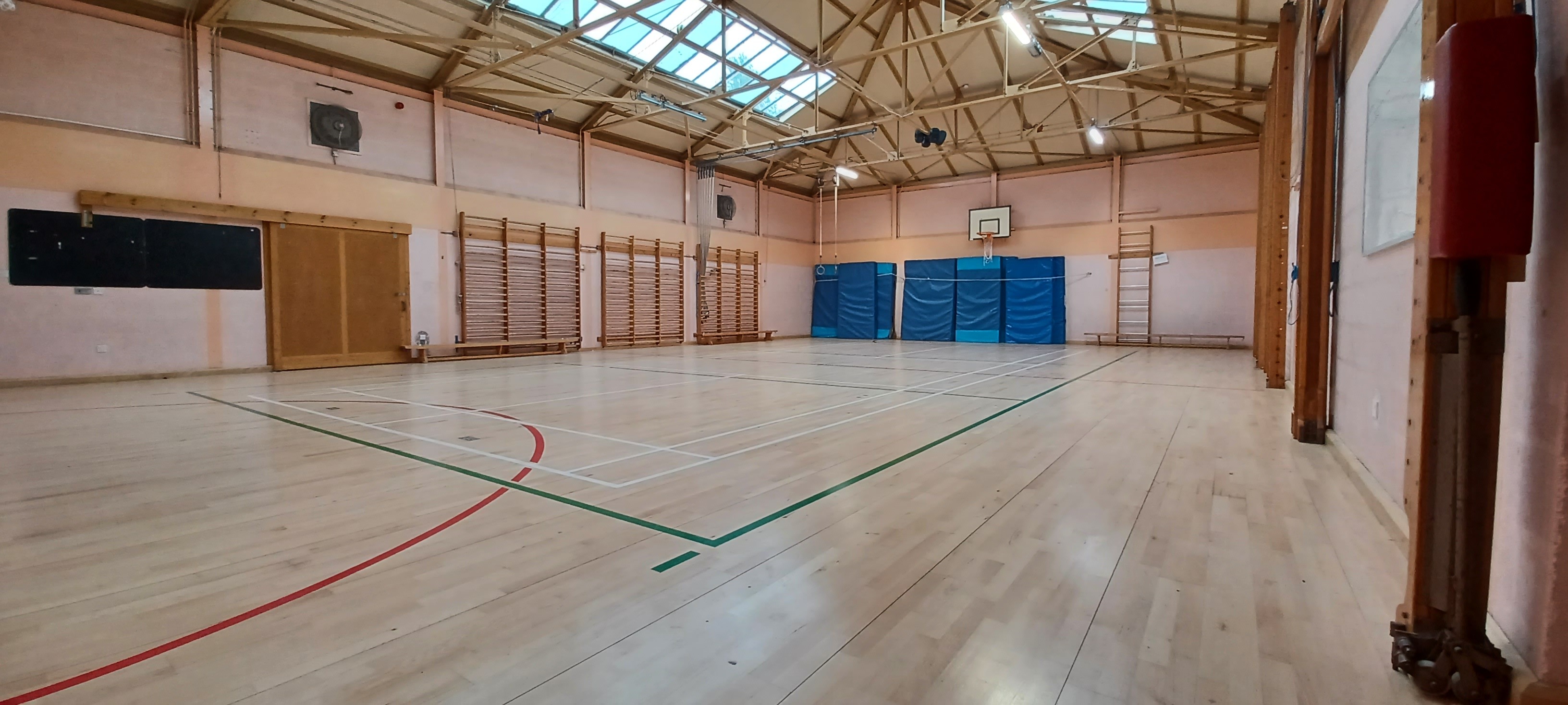 Spacious gymnasium with basketball court markings on the floor, a basketball hoop on the far wall and equipped with apparutus & fitness matts stored against the walls.