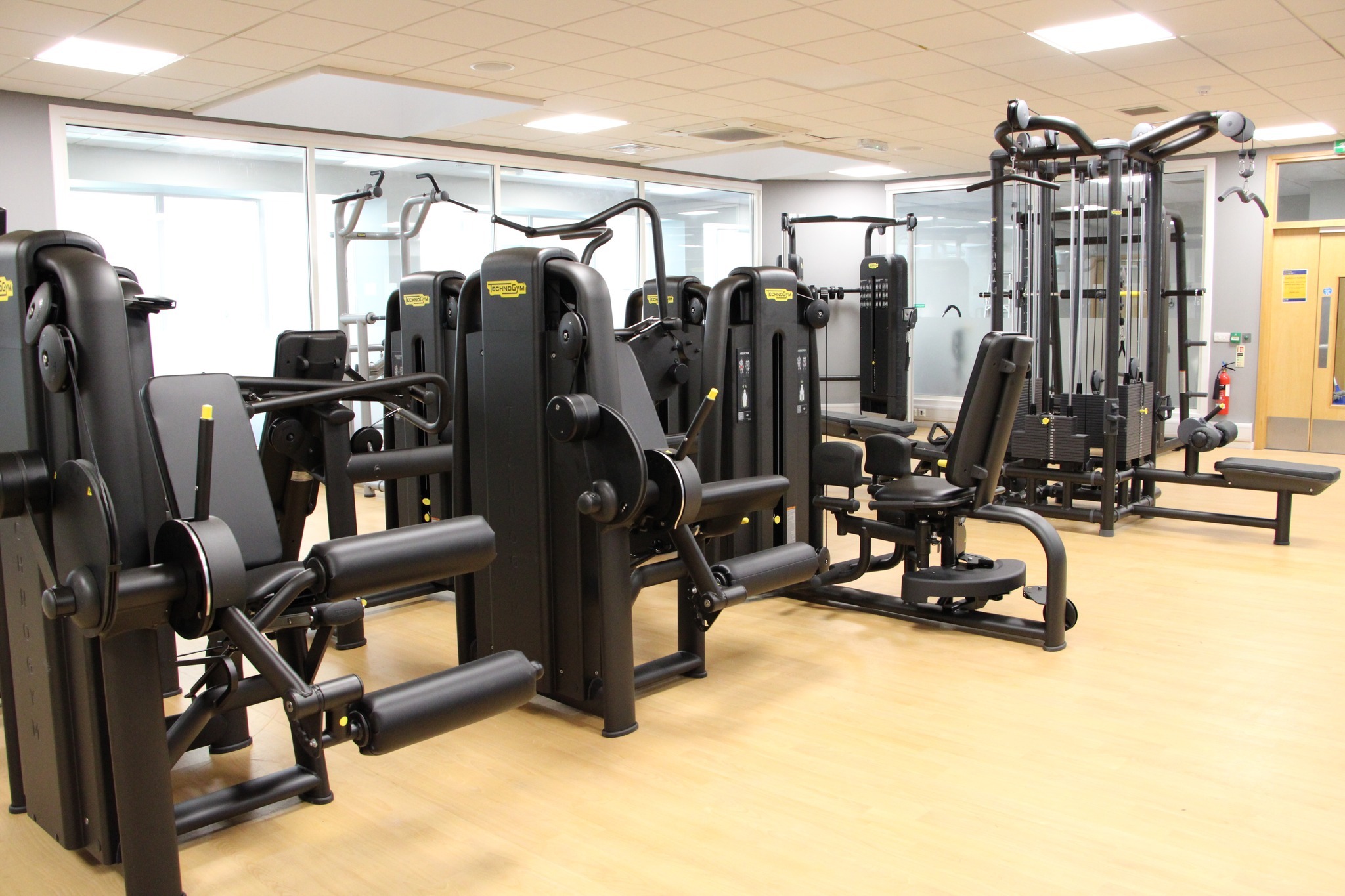 A gym area with several black strength‑training machines arranged on a light wooden floor. The equipment includes leg extension and curl machines, a seated chest press, and a multi-station cable machine. Large windows and mirrors line the back wall, and bright ceiling lights illuminate the space.