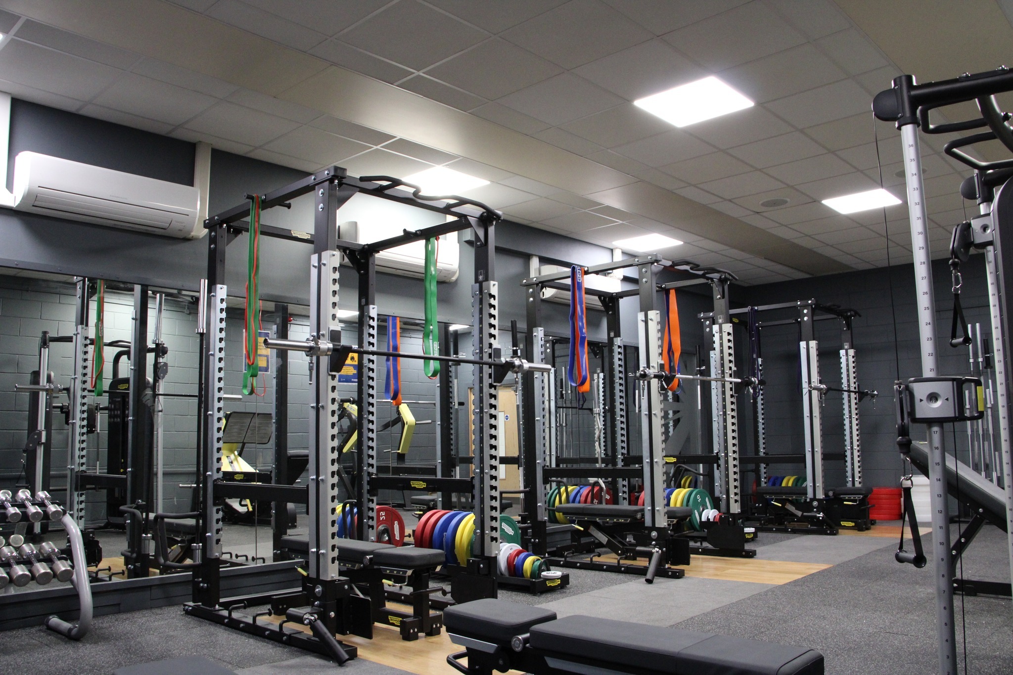 A strength training area with multiple squat racks and lifting platforms, each equipped with colourful weight plates and resistance bands hanging from the frames. Dumbbells are stored on a rack along the mirrored wall to the left, and benches are positioned around the space. The room has grey walls, bright ceiling lights, and a clean, organised layout.