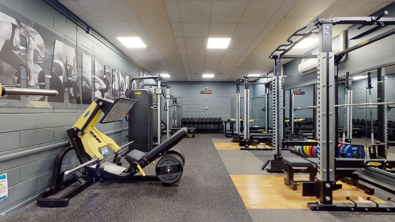 A strength training room with a leg press machine on the left, several weight machines along the back wall, and multiple squat racks with colourful weight plates on the right. The space has rubber and wooden flooring, grey walls with fitness-themed artwork, and bright ceiling lights.