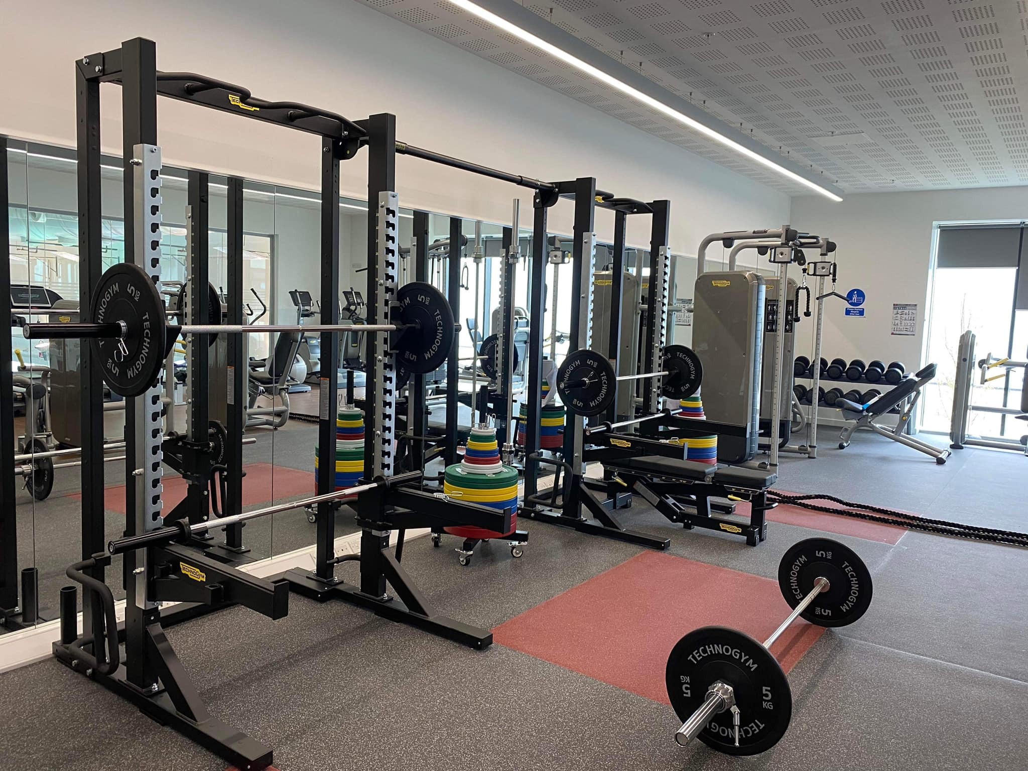 A gym strength area with two squat racks fitted with barbells and weight plates, positioned in front of a mirrored wall. Colourful weight plates are stacked on storage posts between the racks. A cable machine and a rack of dumbbells are visible further back. The room has rubber and red flooring tiles, bright lighting, and a clean, modern layout.