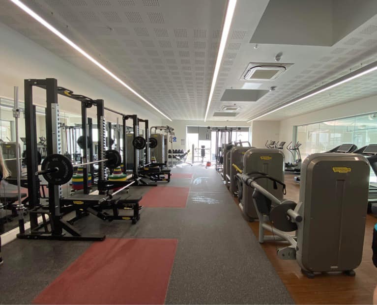 A bright, modern gym with squat racks and weightlifting platforms on the left, each equipped with barbells and colourful weight plates. On the right side are resistance machines arranged in a row. Further back, cardio equipment including treadmills is visible near the windows. The space has rubber and red flooring sections, mirrored walls, and ceiling strip lighting.