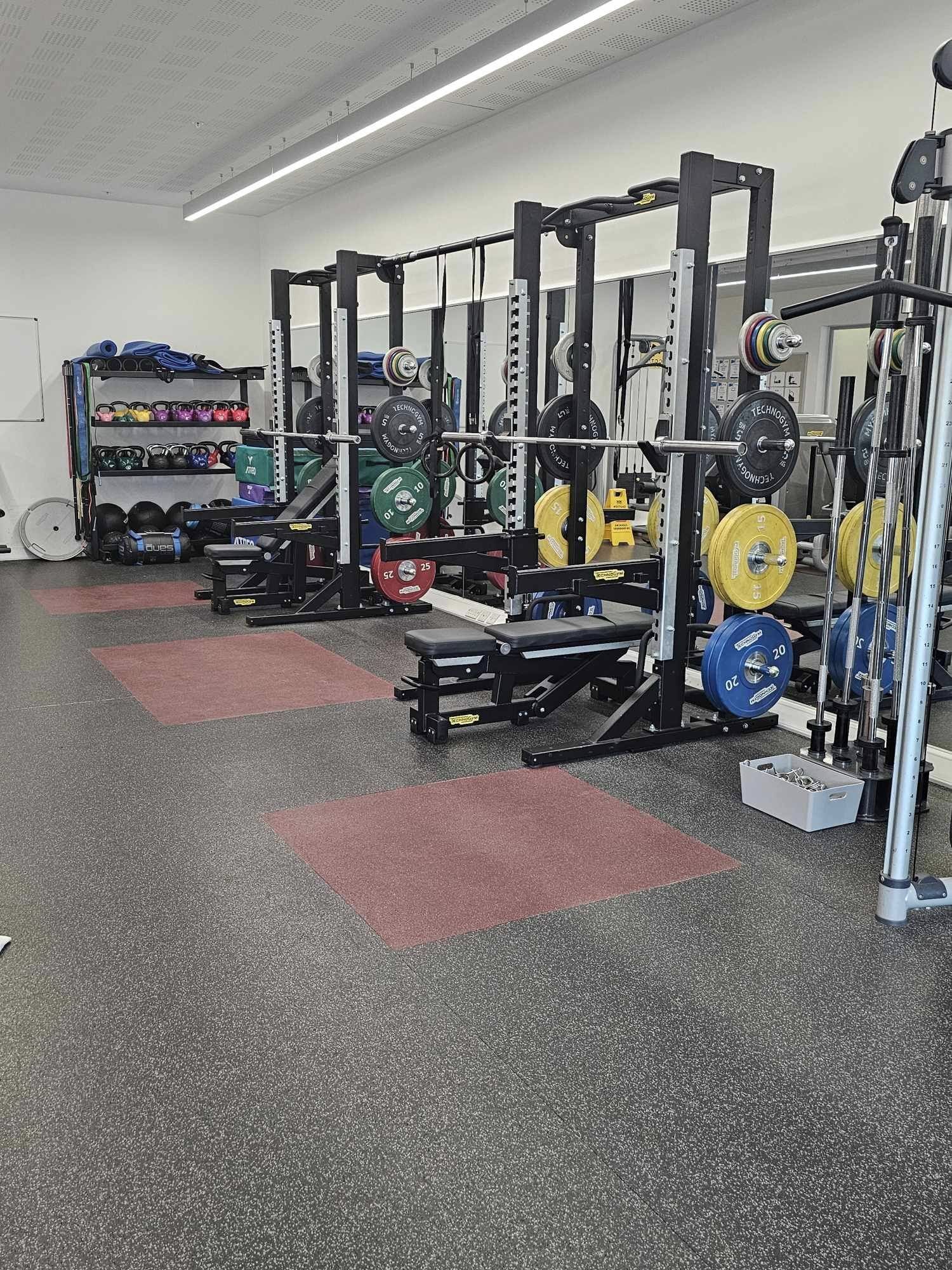A strength and conditioning room with multiple squat racks equipped with barbells and colourful weight plates. Adjustable benches sit beneath the racks, and a storage shelf in the background holds kettlebells, medicine balls, and other training equipment. The room has rubber flooring with red lifting platforms and bright overhead lighting