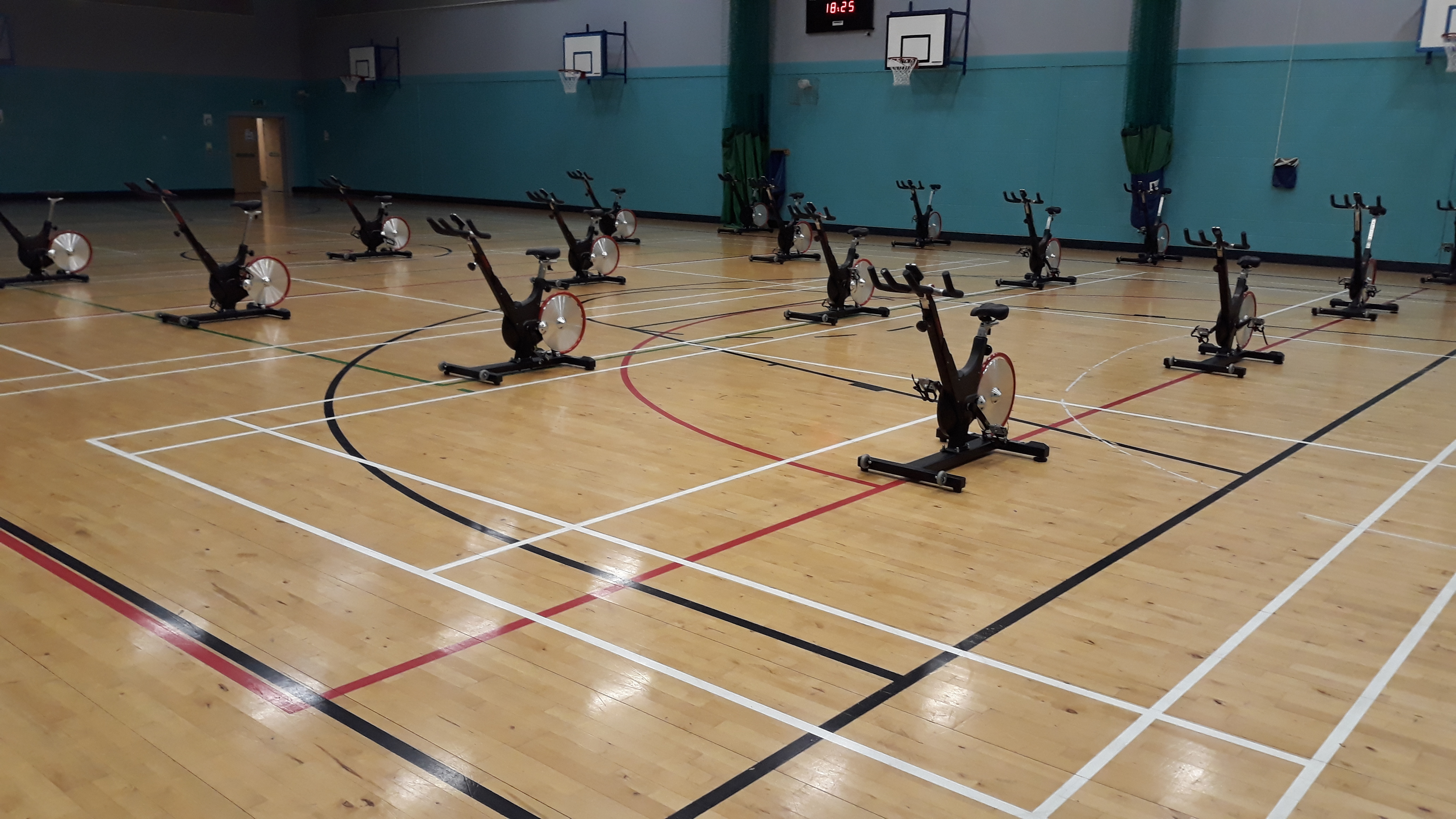 A sports hall with spaced‑out indoor cycling bikes arranged across the wooden floor.
