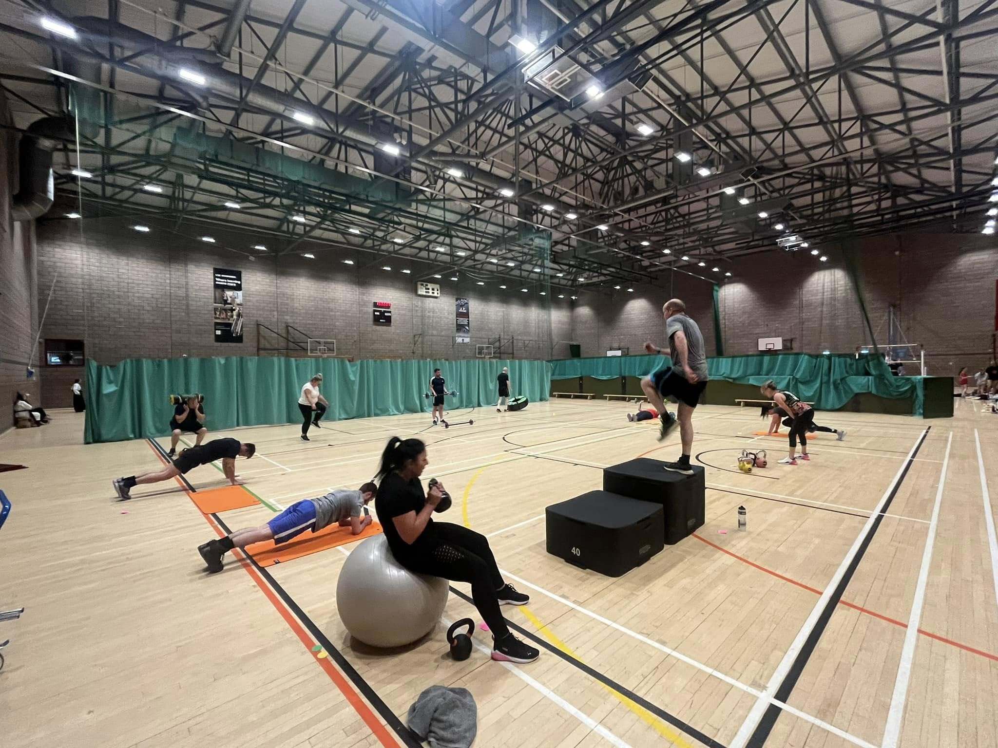 People taking part in a circuit‑style fitness class in a large sports hall, using equipment such as steps, mats, weights and an exercise ball across different workout stations.