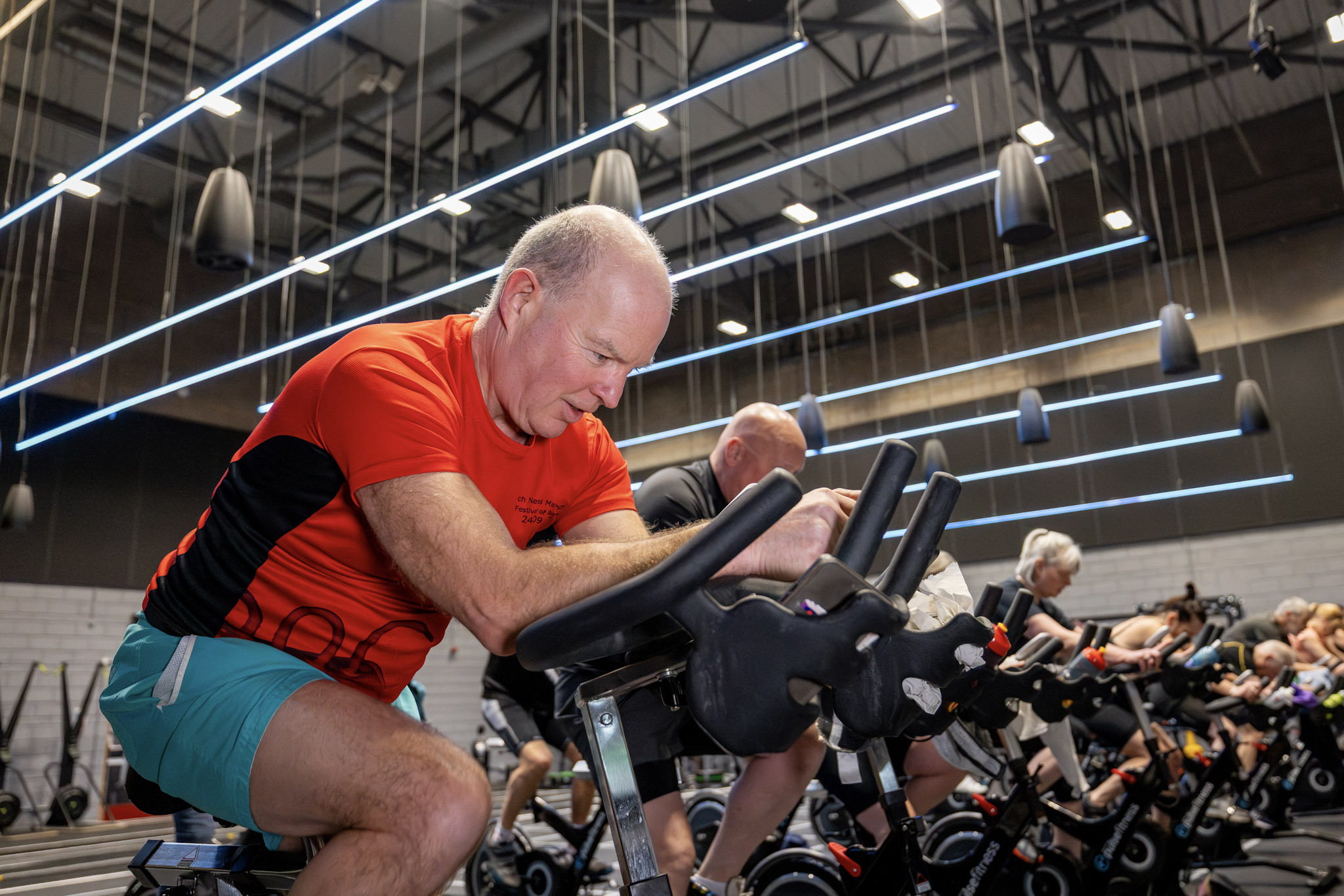 A group of people taking part in an indoor cycling class, riding stationary bikes in a large, modern fitness studio.