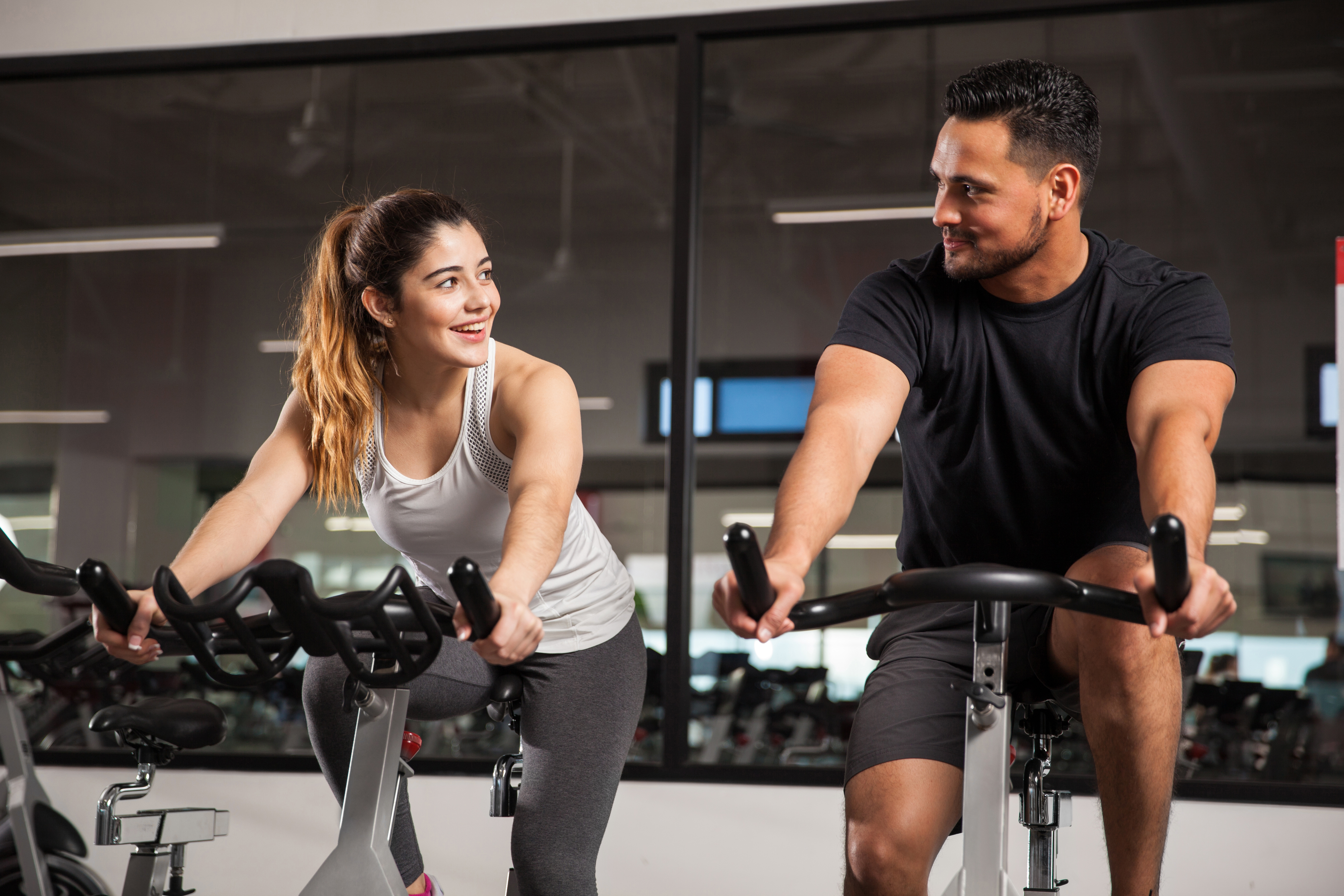 Two people riding stationary exercise bikes in a gym, leaning forward on the handlebars during a cycling workout.